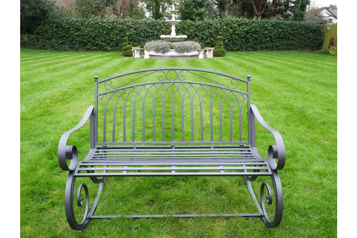 Black metal garden bench with decorative scroll arms on lawn facing fountain