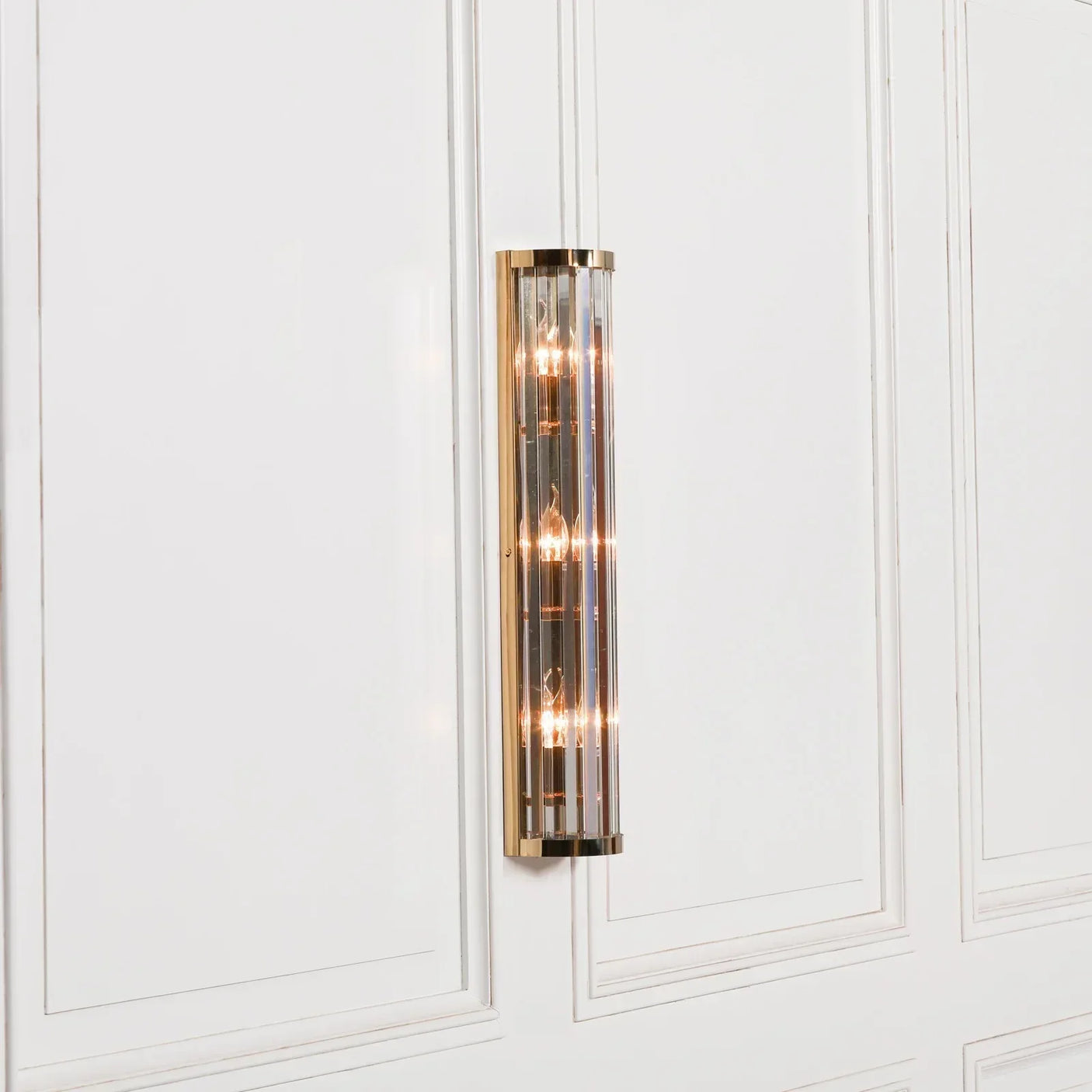 Brass wall light with vertical glass panels mounted on white panelled wall