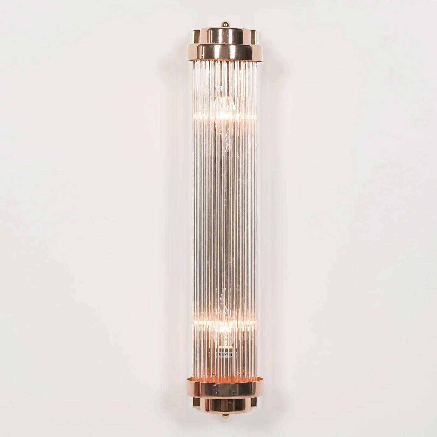 Vertical glass ribbed wall light with gold metal ends, illuminated against white wall