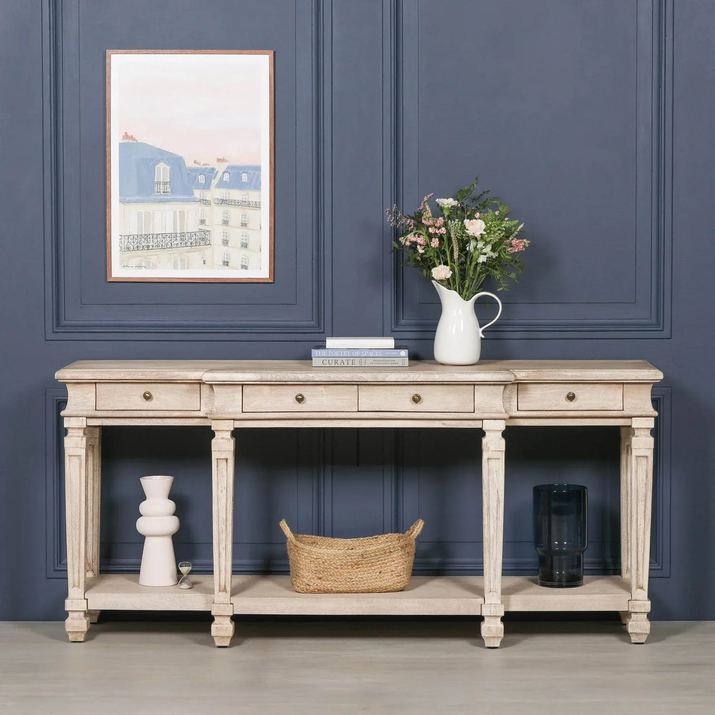 Light wood console table with drawers, shelf, books, vase of flowers, and basket in hallway