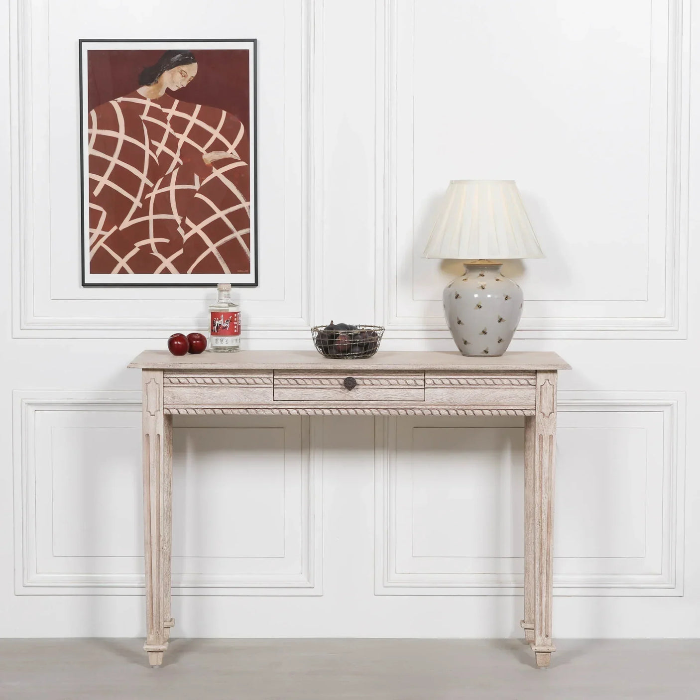 Light wood console table with decorative front, lamp, wire basket, and wall art in hallway