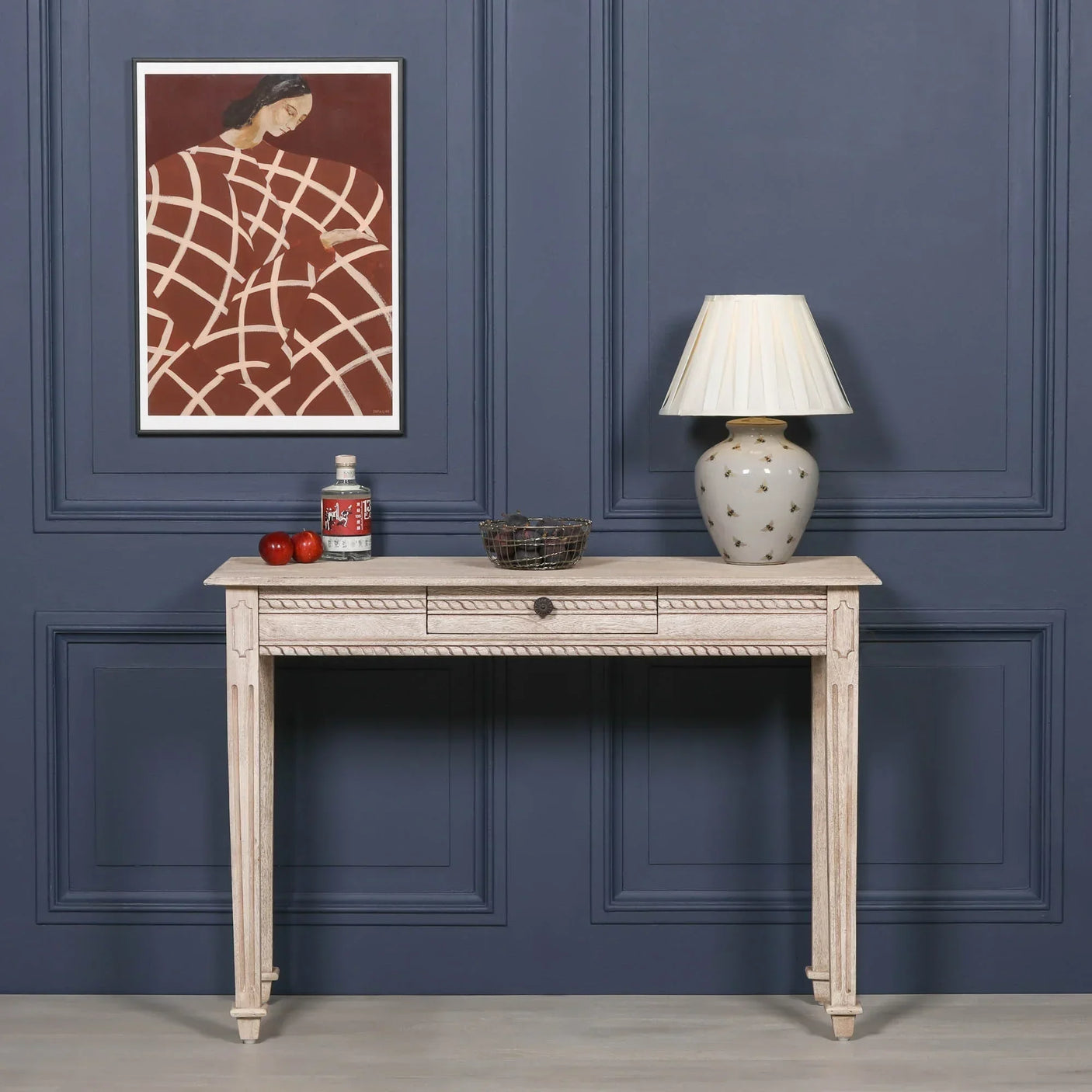 Light wood console table with carved detailing, lamp, and decor against blue panel wall