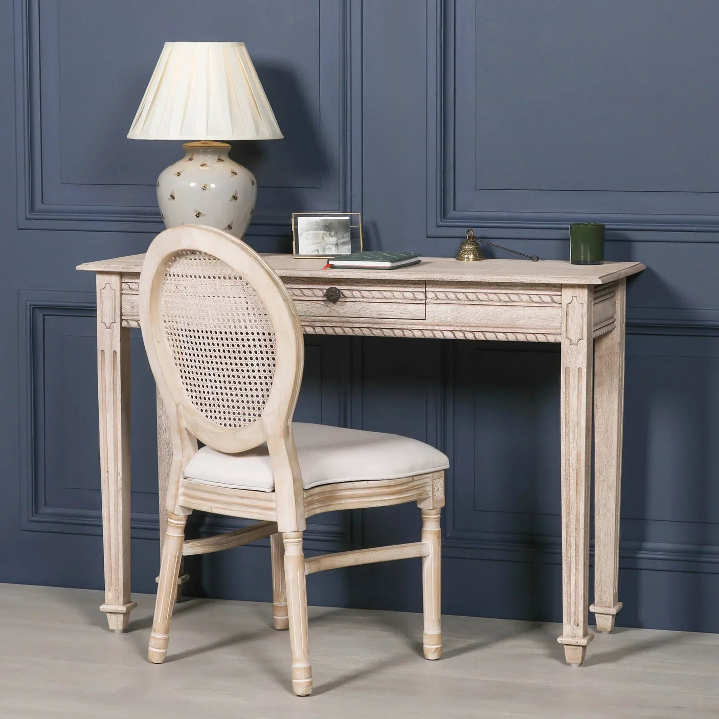 Whitewashed wooden writing desk with cane back chair and lamp against blue panel wall