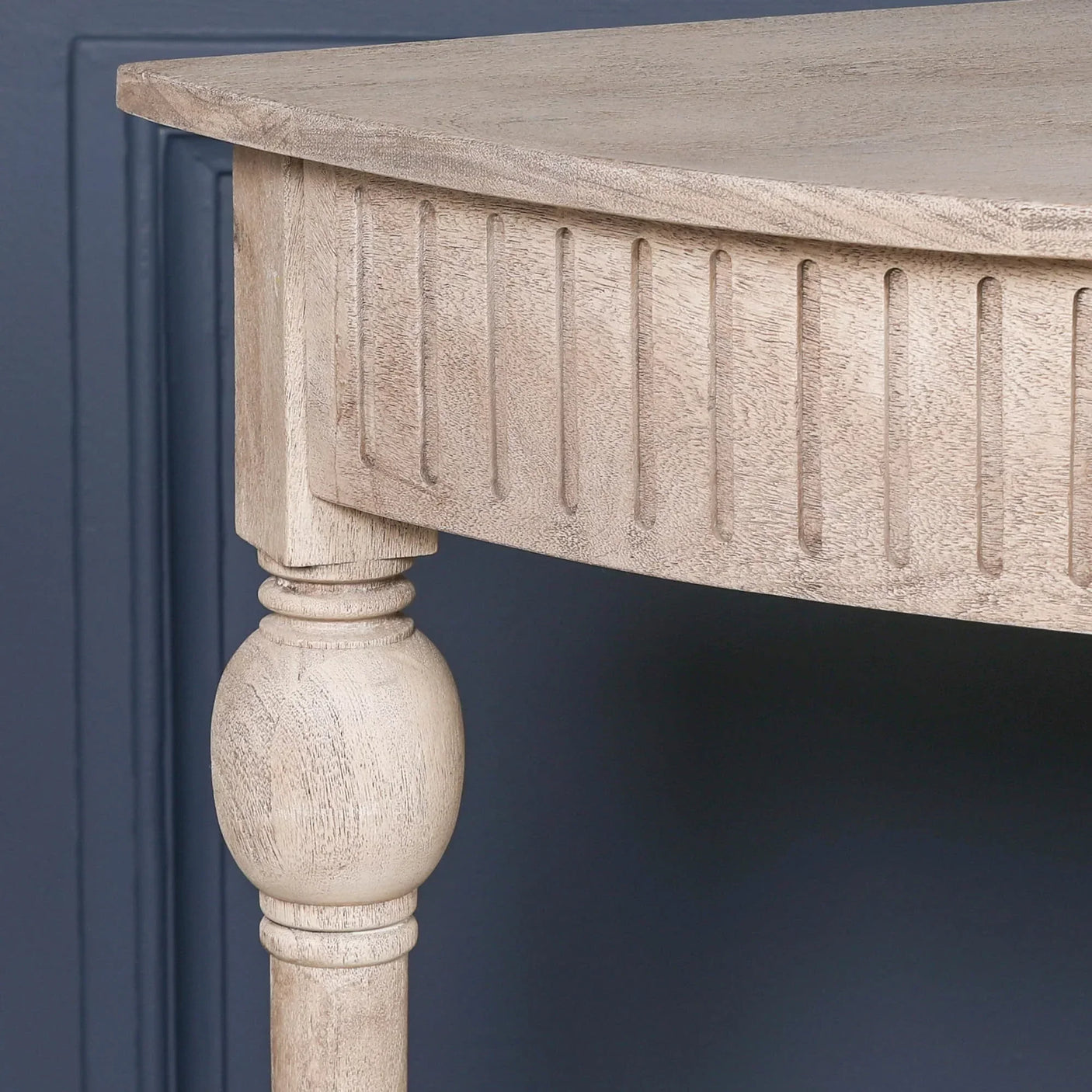 Light wood console table corner with carved groove detail and turned leg against navy wall