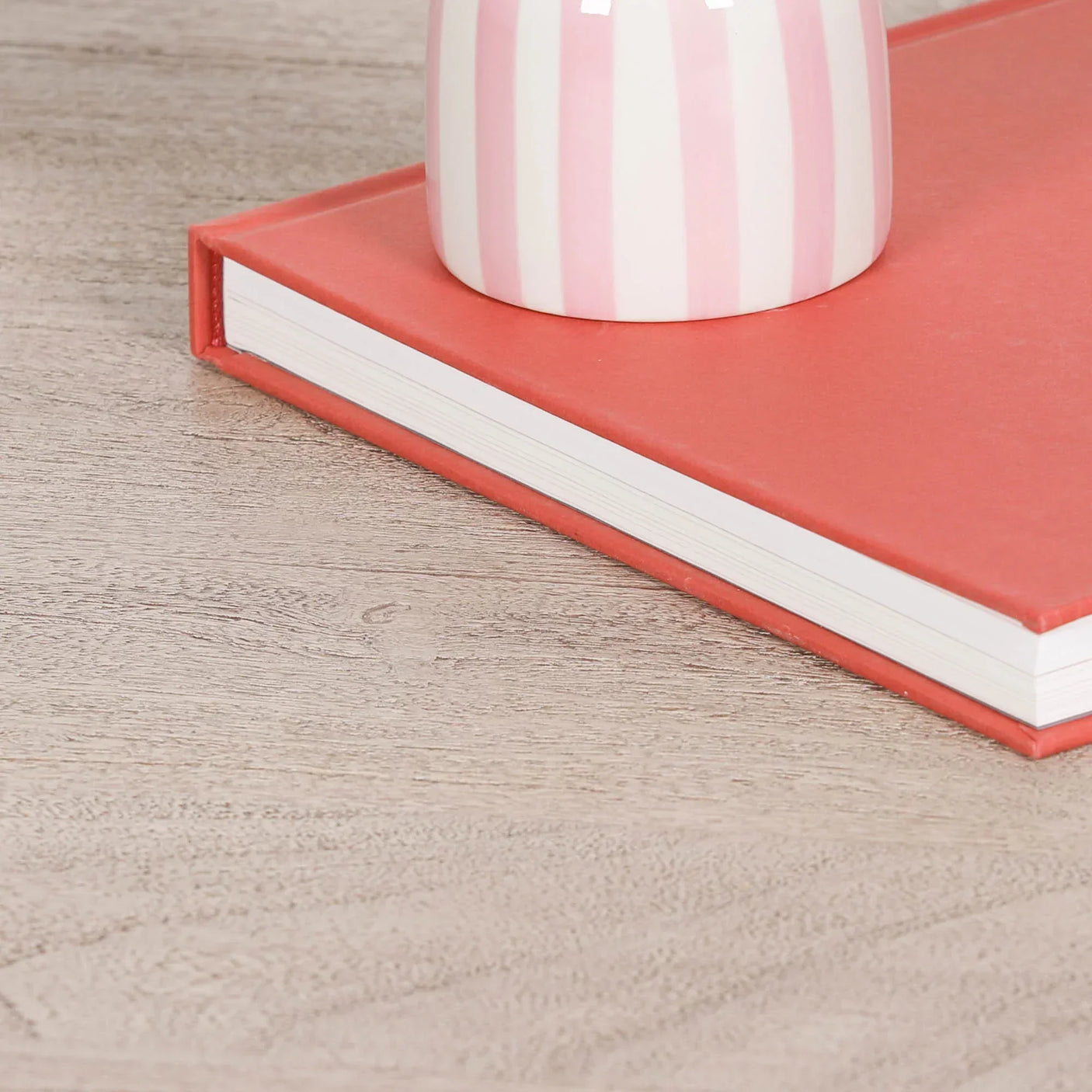 Wood effect table surface with coral hardcover book and striped ceramic vase on top
