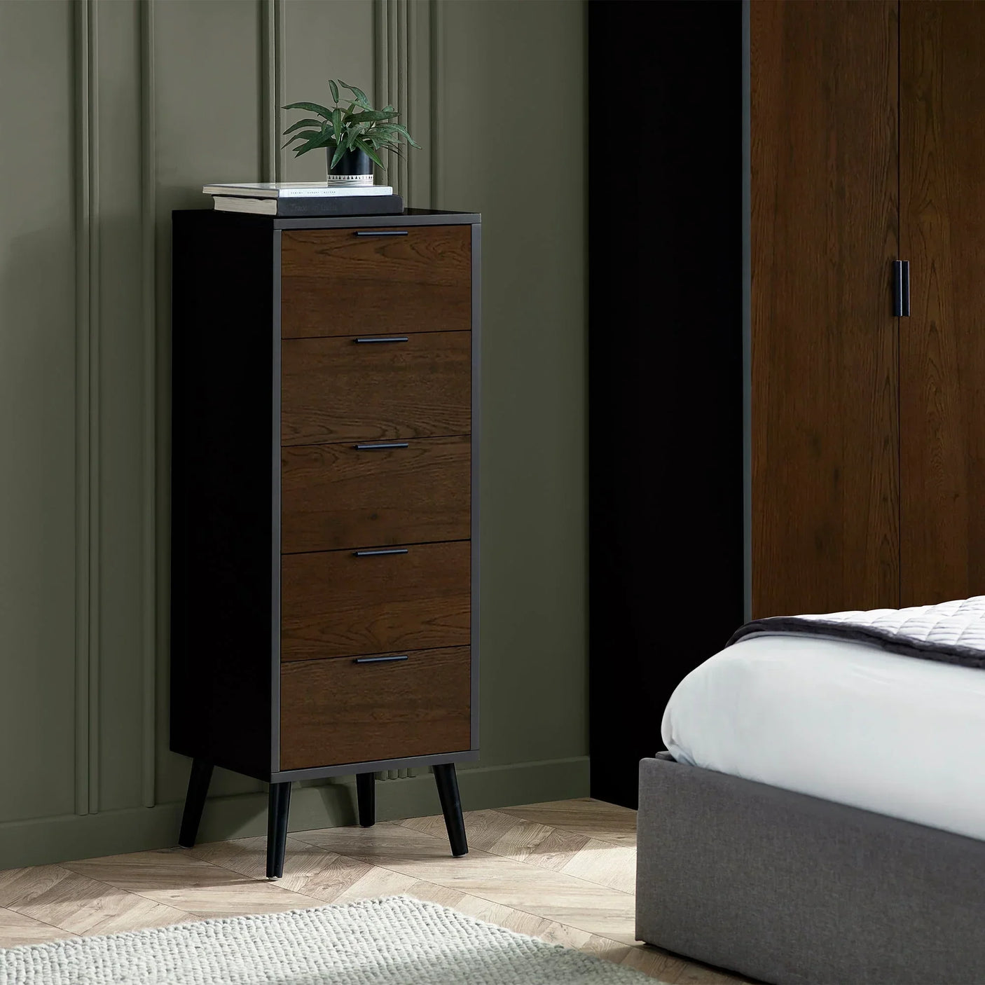 Walnut black five drawer chest with angled legs beside bed in modern bedroom