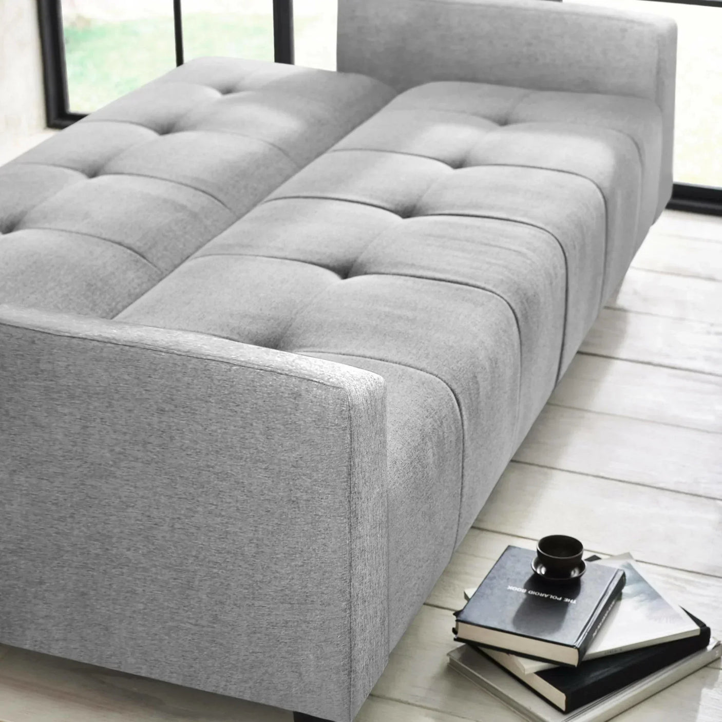 Grey upholstered sofa bed in open position with books and coffee cup on wooden floor