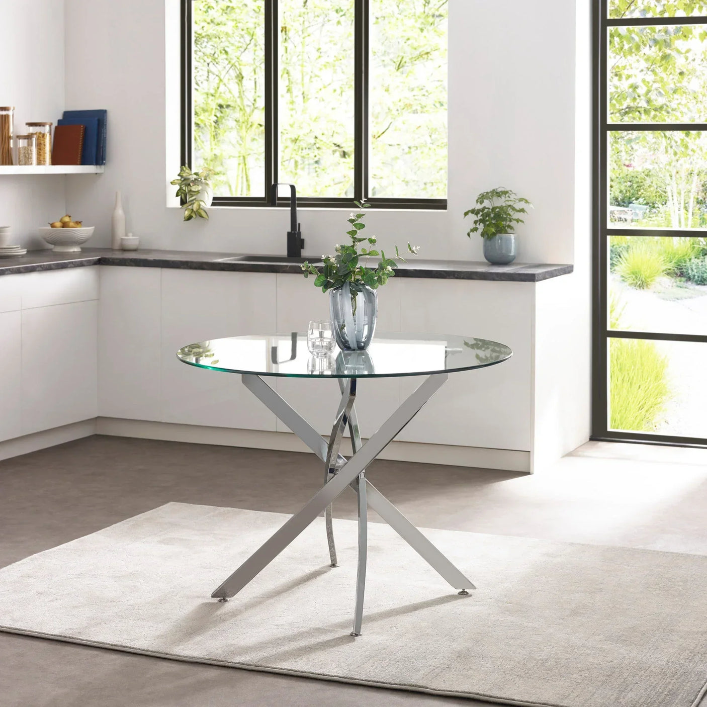 Round glass dining table with chrome legs on a rug in modern kitchen with large windows