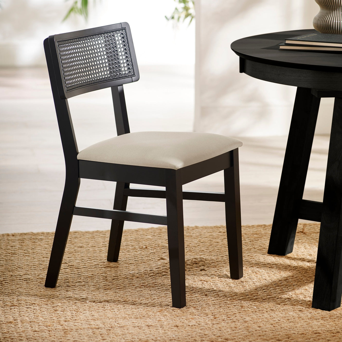 Black dining chair with natural fabric seat and rattan backrest angled left on woven rug beside round black table