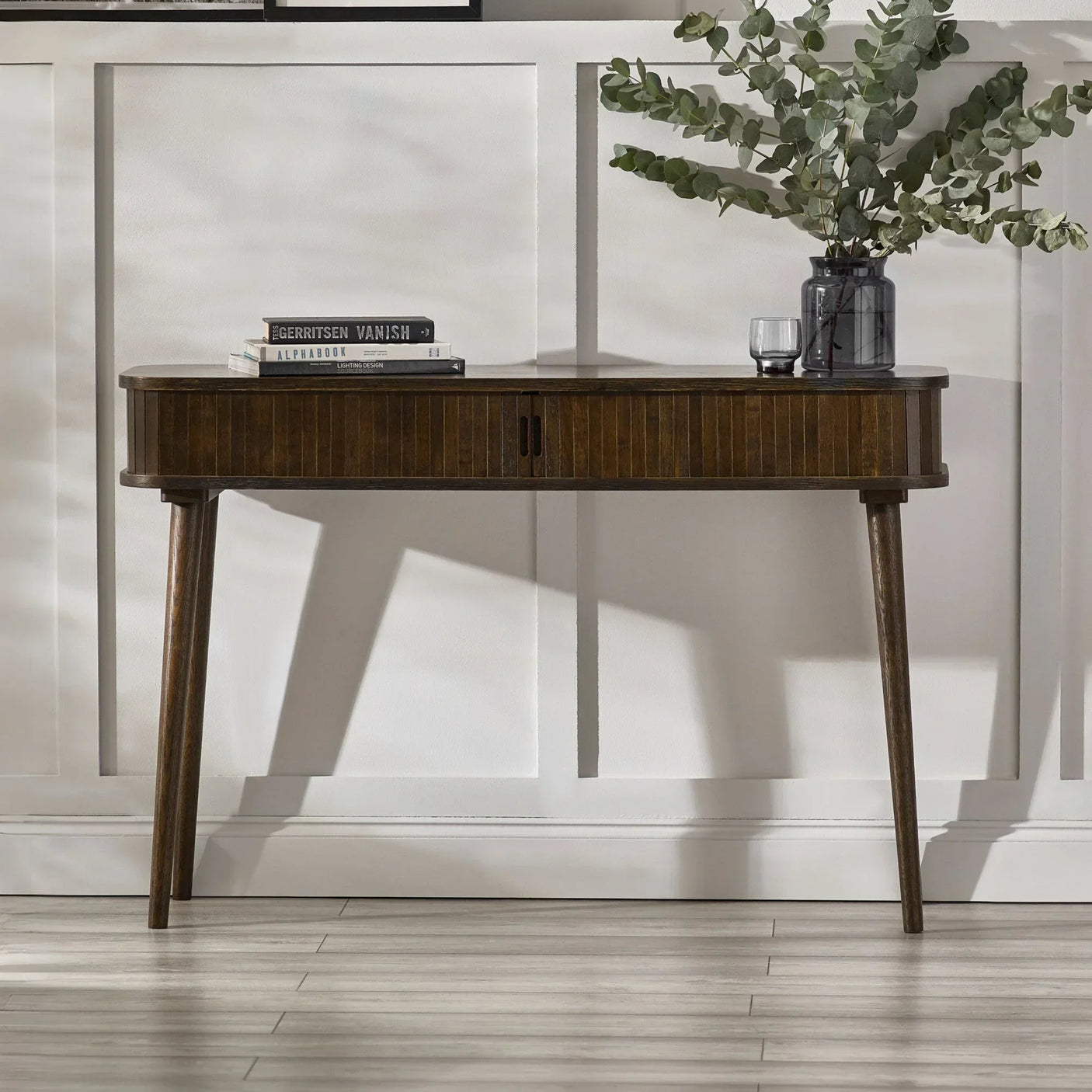 Walnut console table with slim legs, books, glass vase and greenery against white wall panel