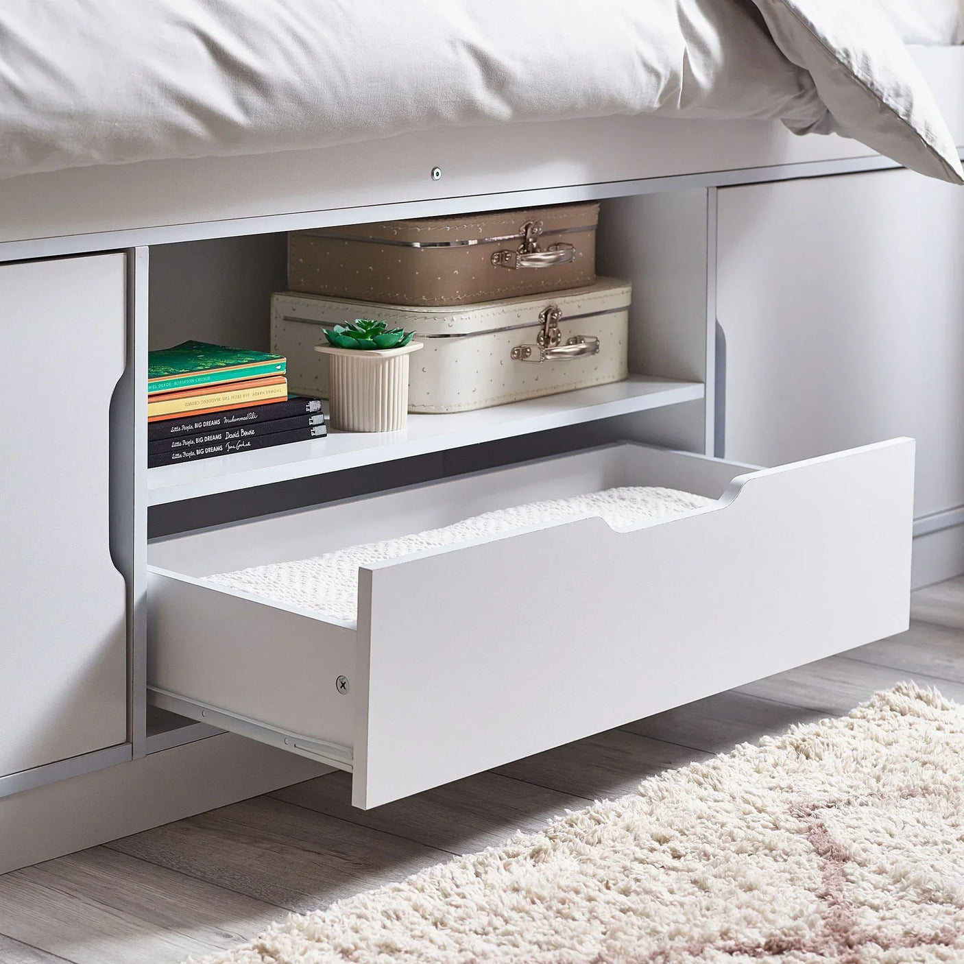 White low sleeper bed with open storage drawer, shelves, and books on wooden floor