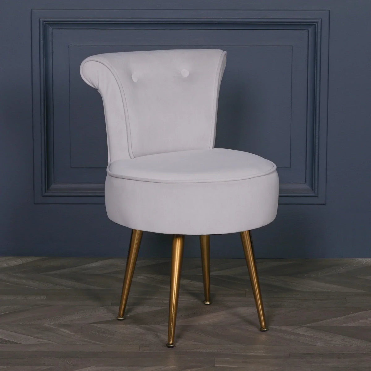 Grey velvet accent chair with gold legs against a dark blue wall and herringbone floor