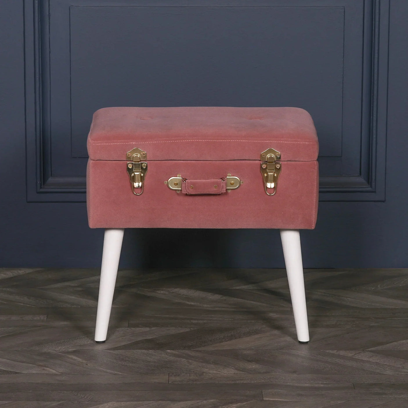 Blush pink velvet ottoman with suitcase design and white legs against blue wall