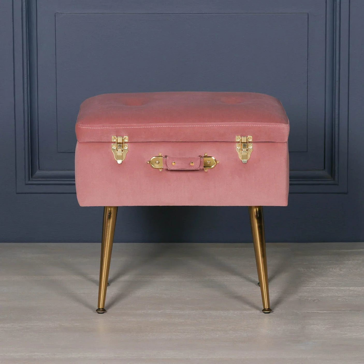 Pink velvet ottoman with gold legs and suitcase-style handle against navy wall