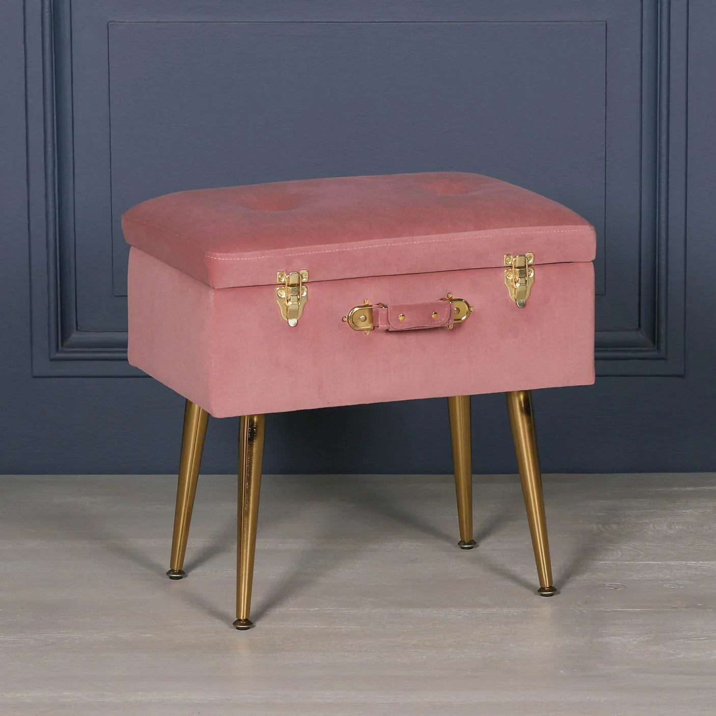 Pink velvet storage ottoman with gold metal legs and brass latches against dark wall