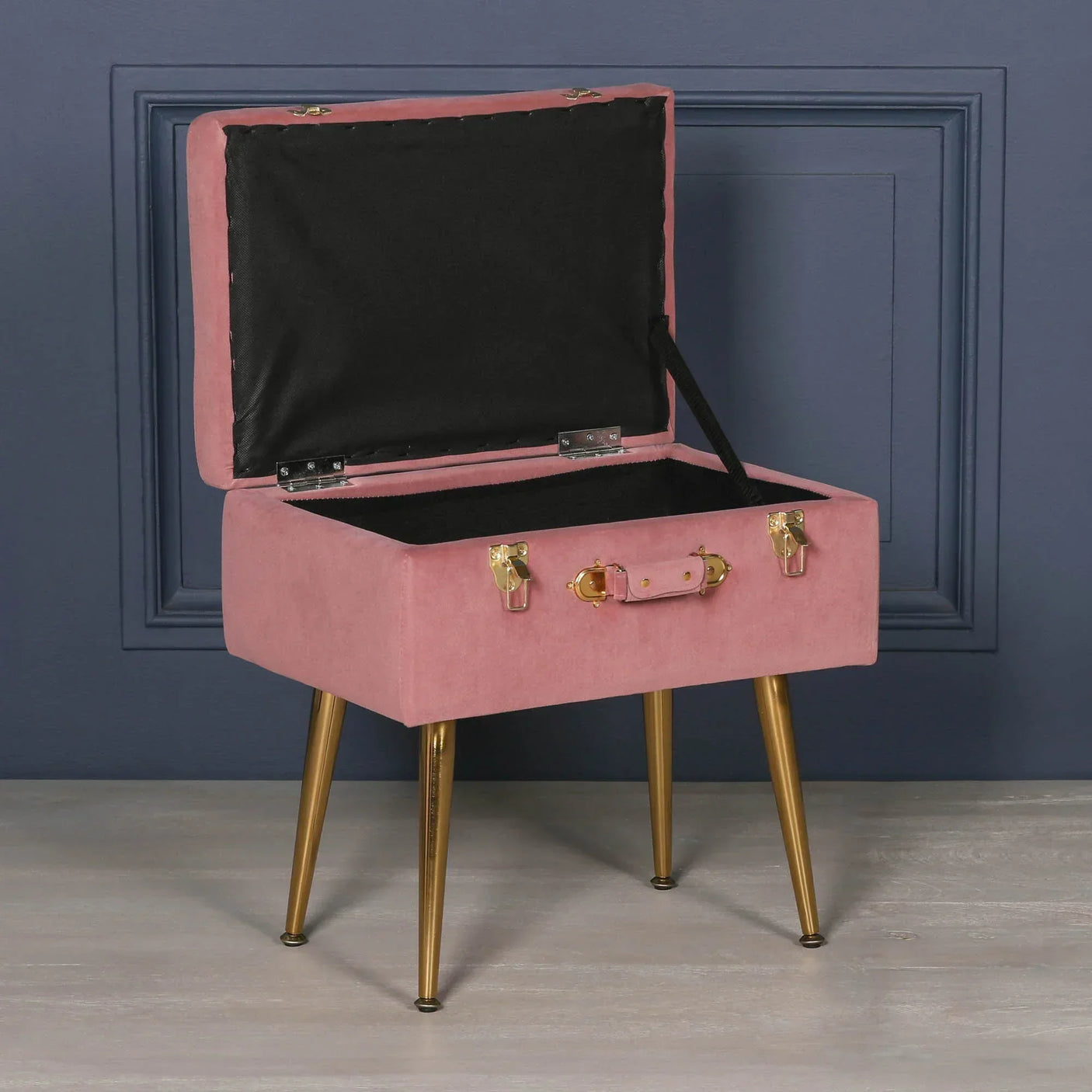Pink velvet storage stool with gold legs and open lid against dark blue wall