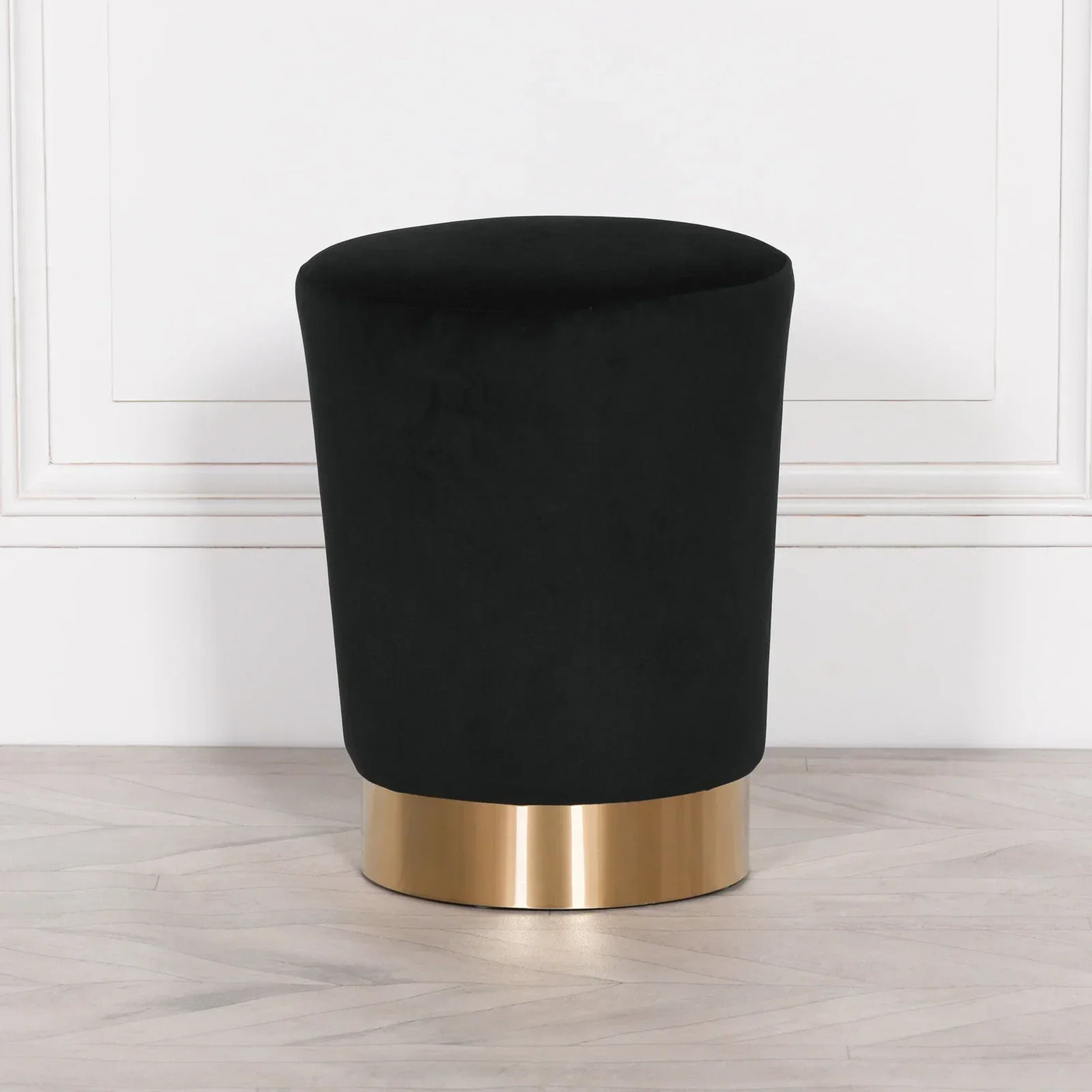 Black velvet stool with gold metal base on light wooden floor against white panel wall