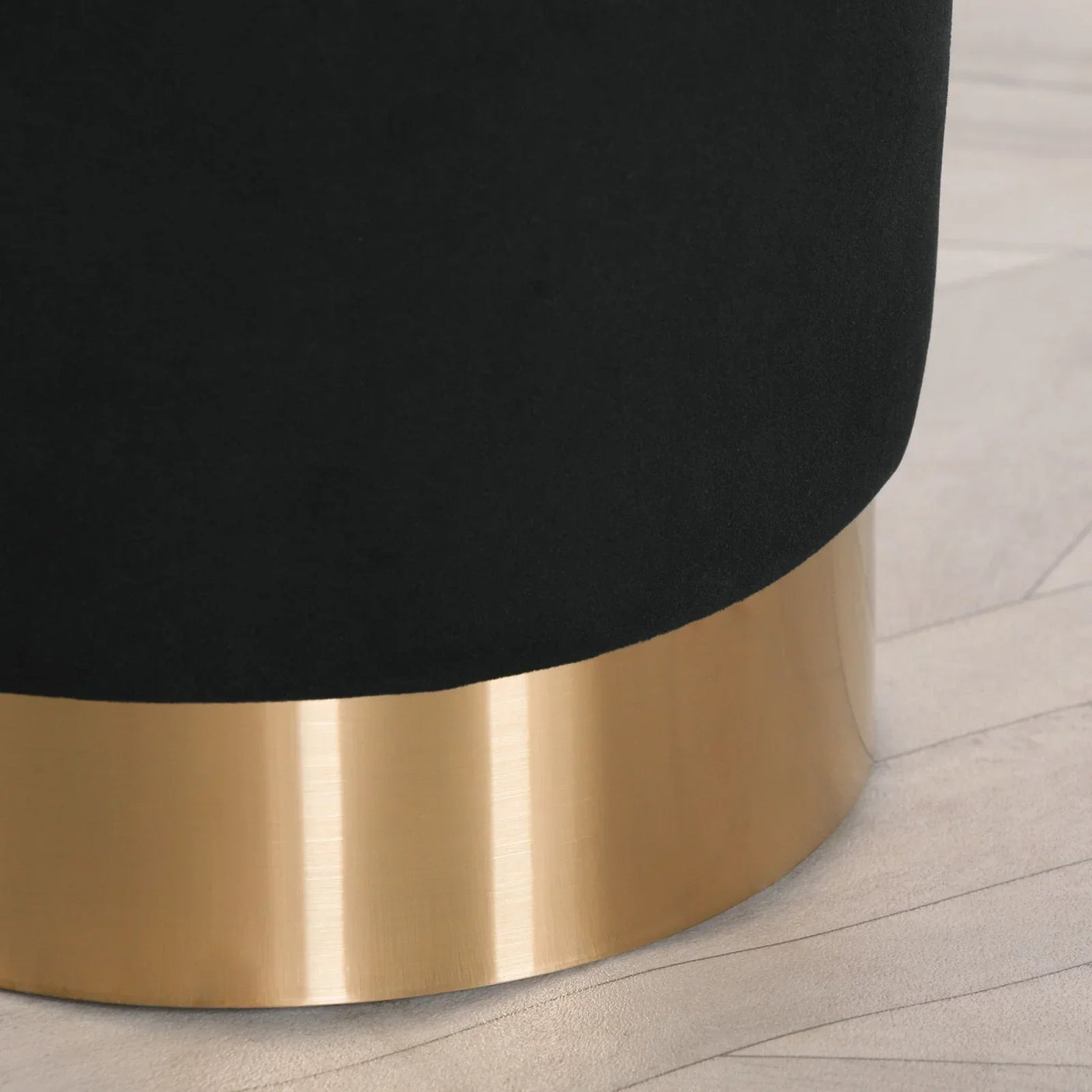 Black velvet ottoman with gold metal base on light patterned floor, close-up view
