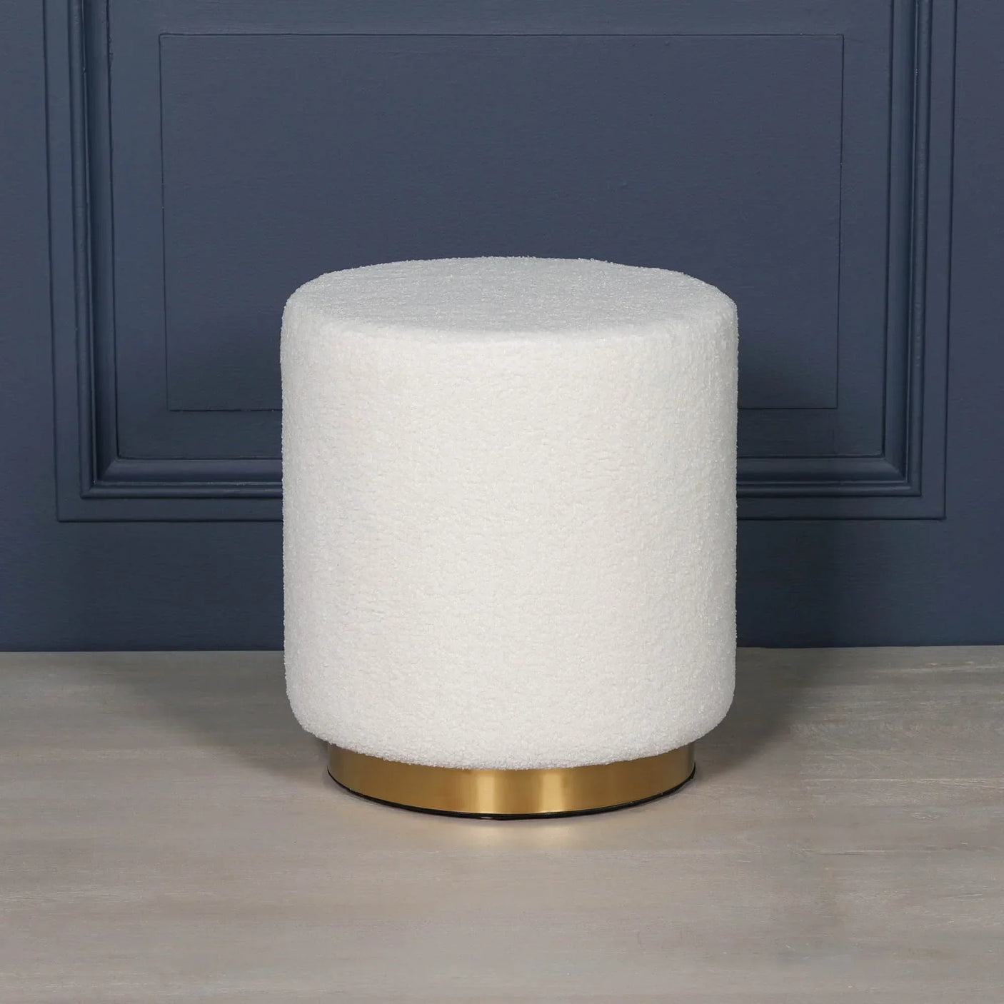 Round white boucle ottoman with gold base on wooden floor against navy wall