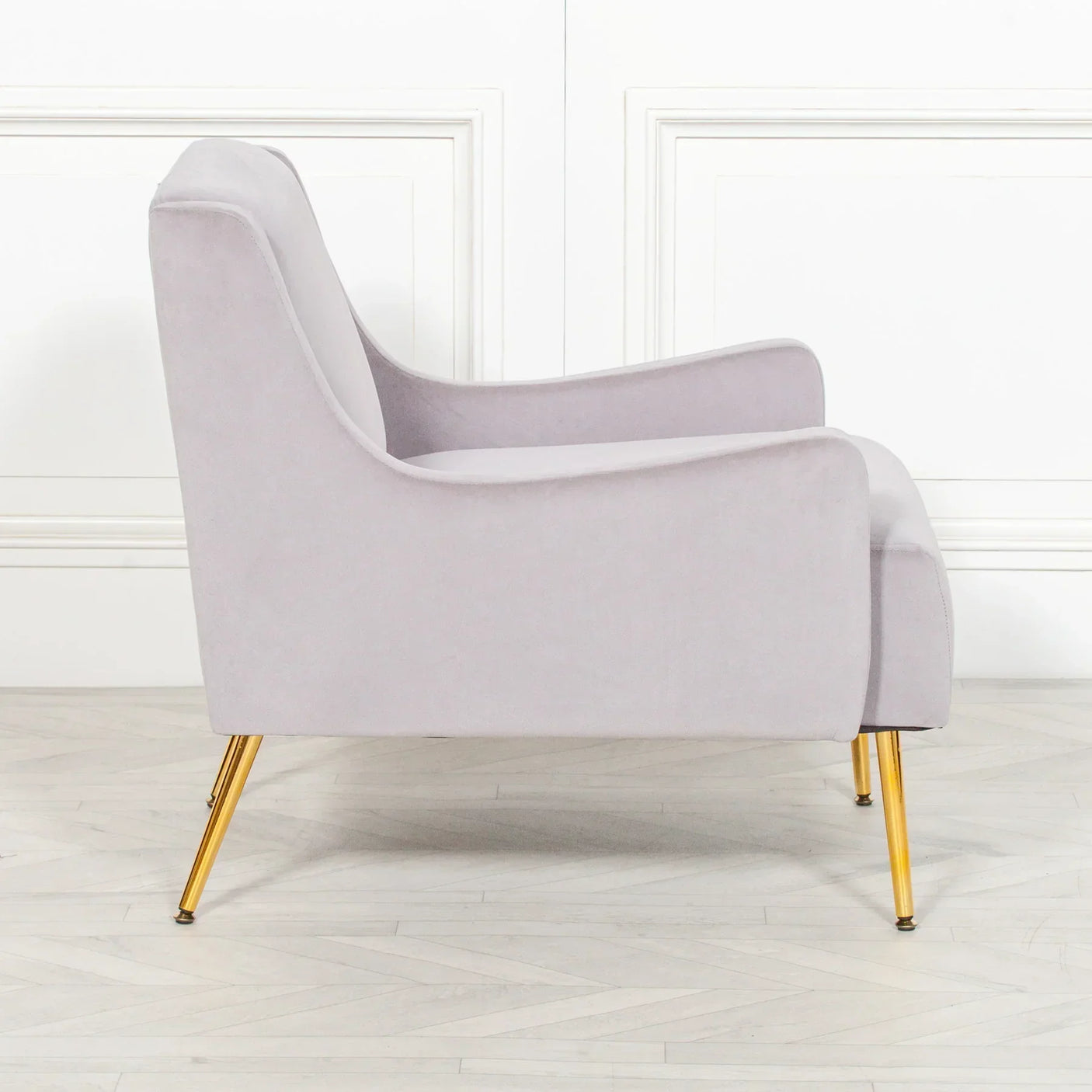 Light grey velvet armchair with gold metal legs, side view on wooden floor