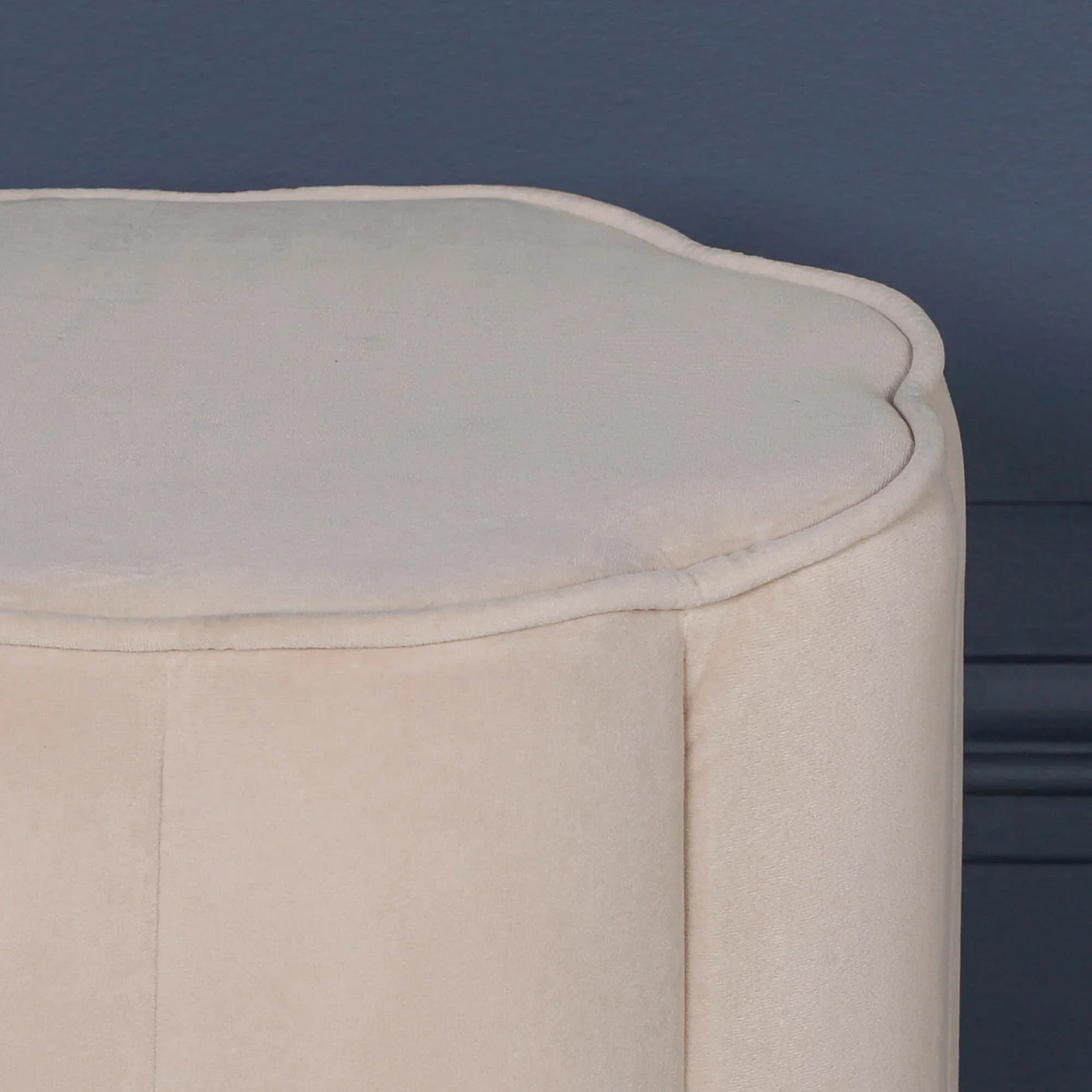 Beige velvet ottoman with scalloped top edge against blue wall