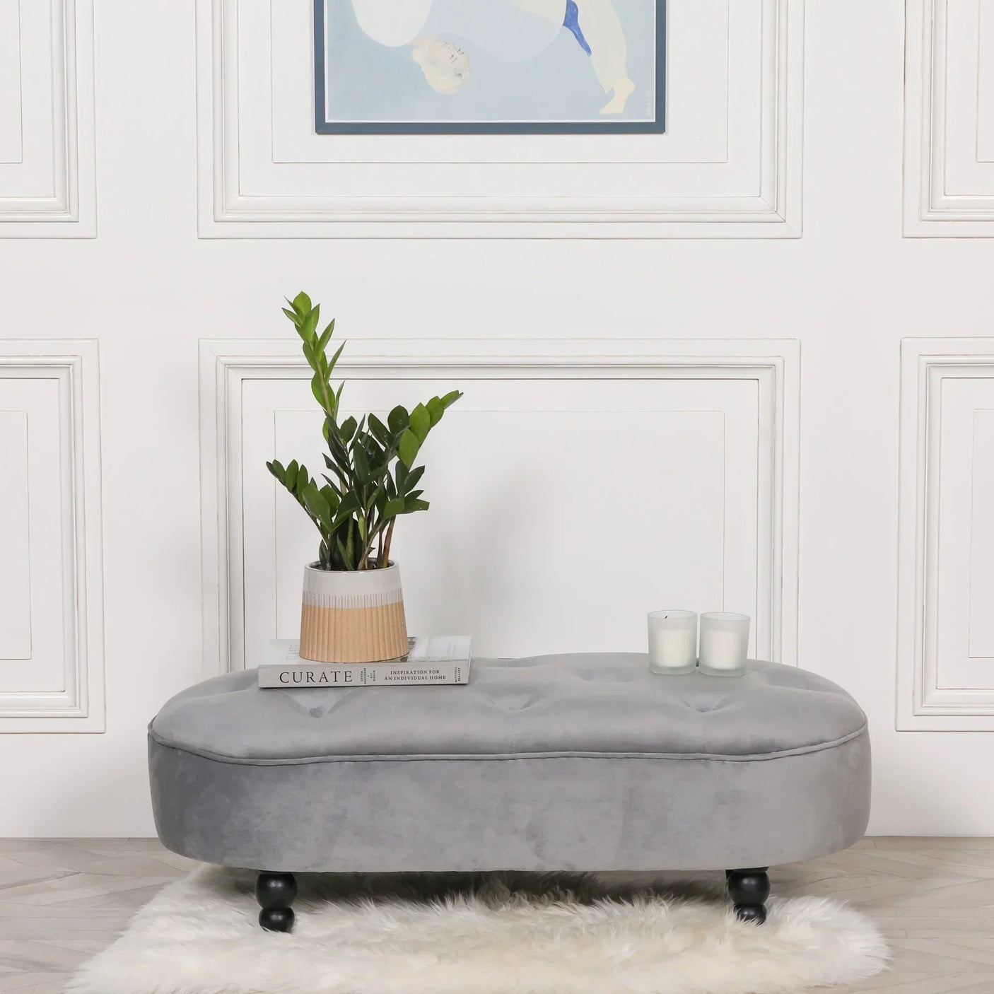Grey velvet ottoman bench with black wooden legs, plant, candles and books on top