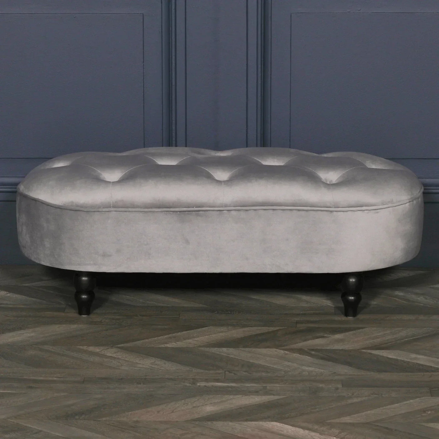 Grey velvet ottoman bench with button-tufted top and dark wooden legs on parquet floor