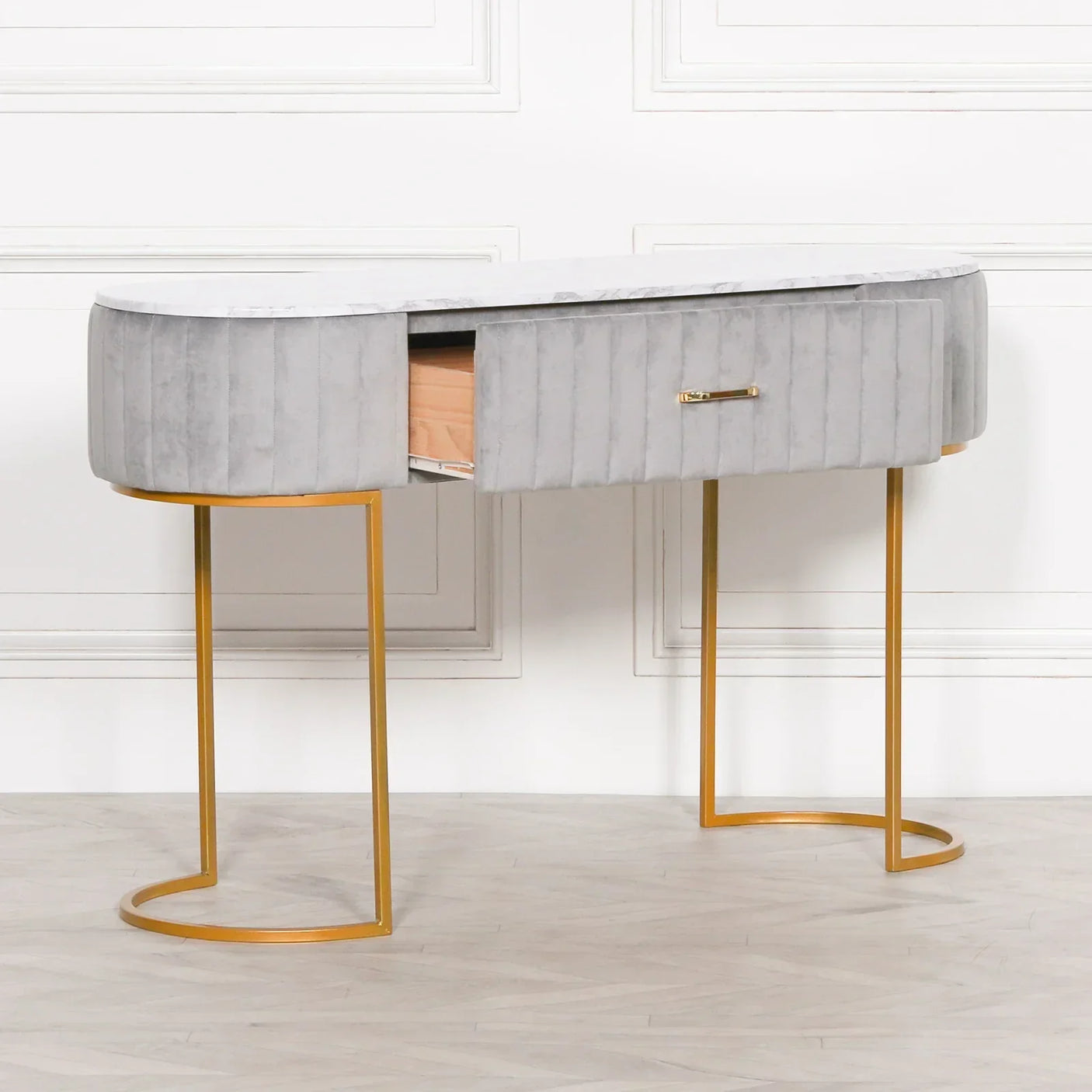 Grey velvet console table with gold legs and open drawer against white paneled wall