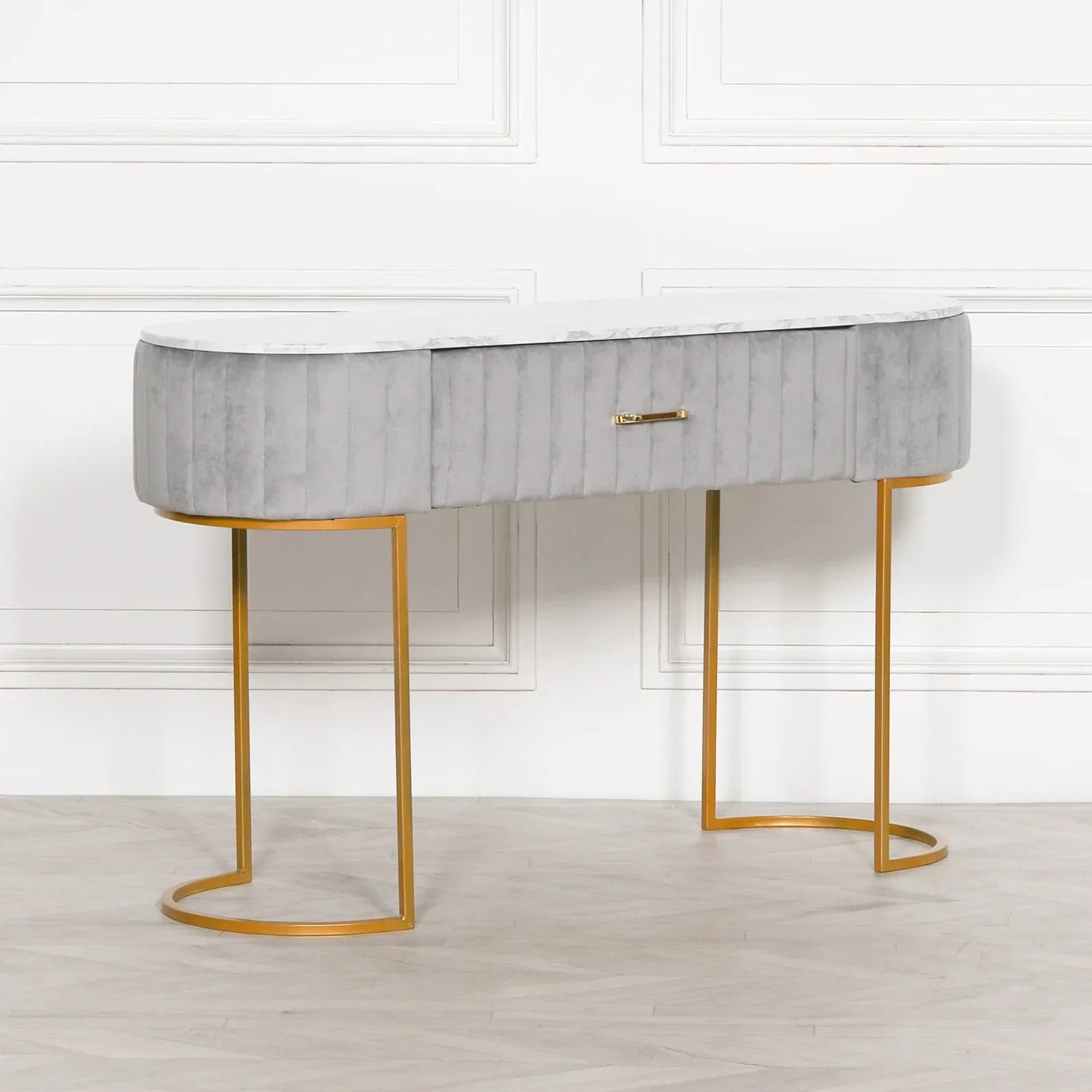 Grey velvet console table with gold metal legs and single drawer, placed against white wall