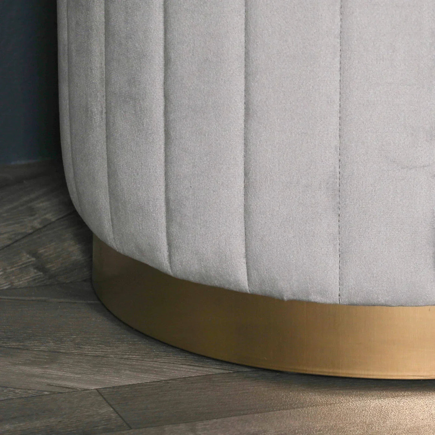 Close view of grey velvet ottoman with vertical stitching and gold metal base on wood floor