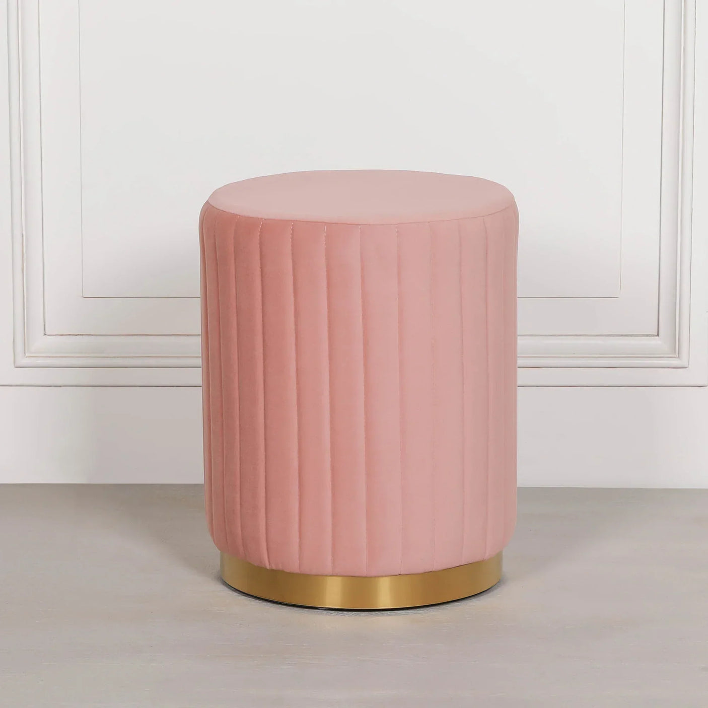 Pink velvet ottoman stool with vertical stitching and gold base against white wall