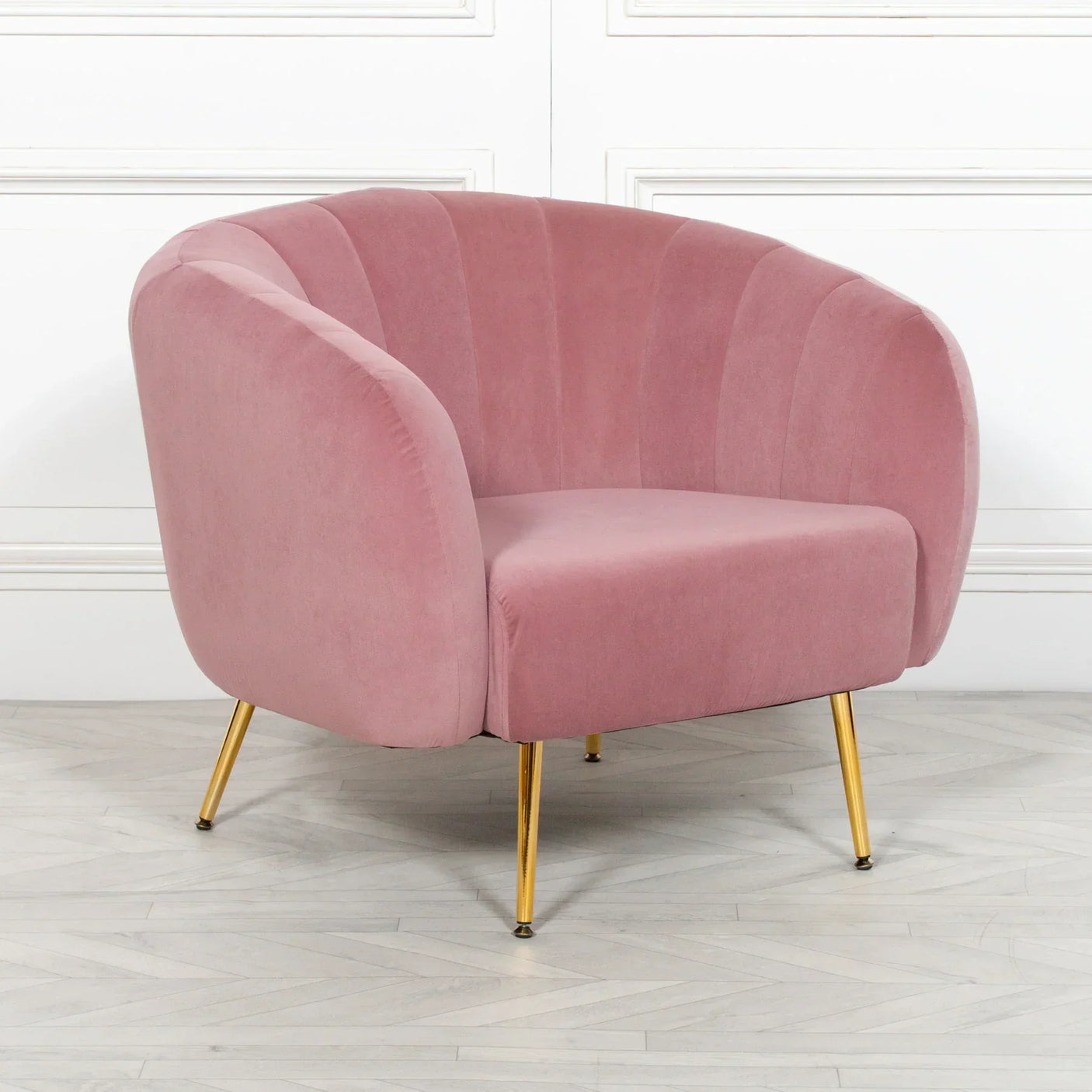 Pink velvet accent chair with gold legs on herringbone wood floor against white paneled wall