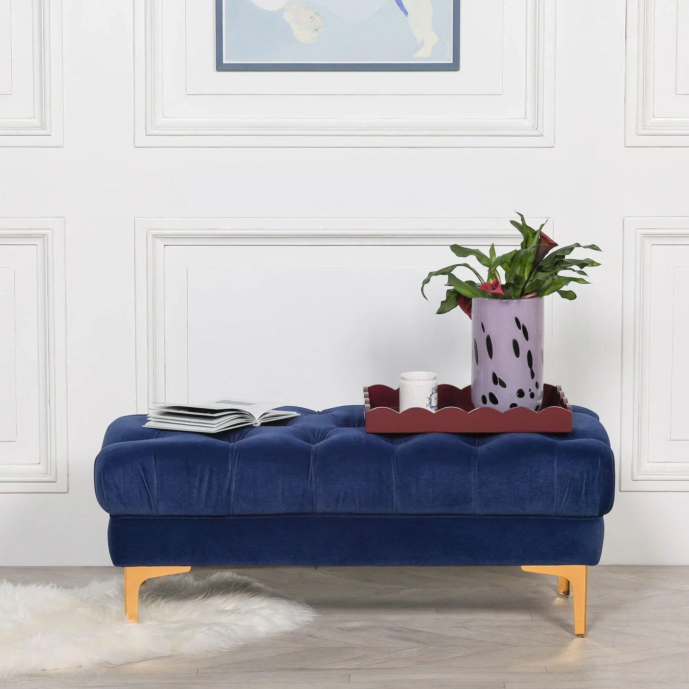 Blue velvet ottoman bench with gold legs, tray, plant and open book against white wall