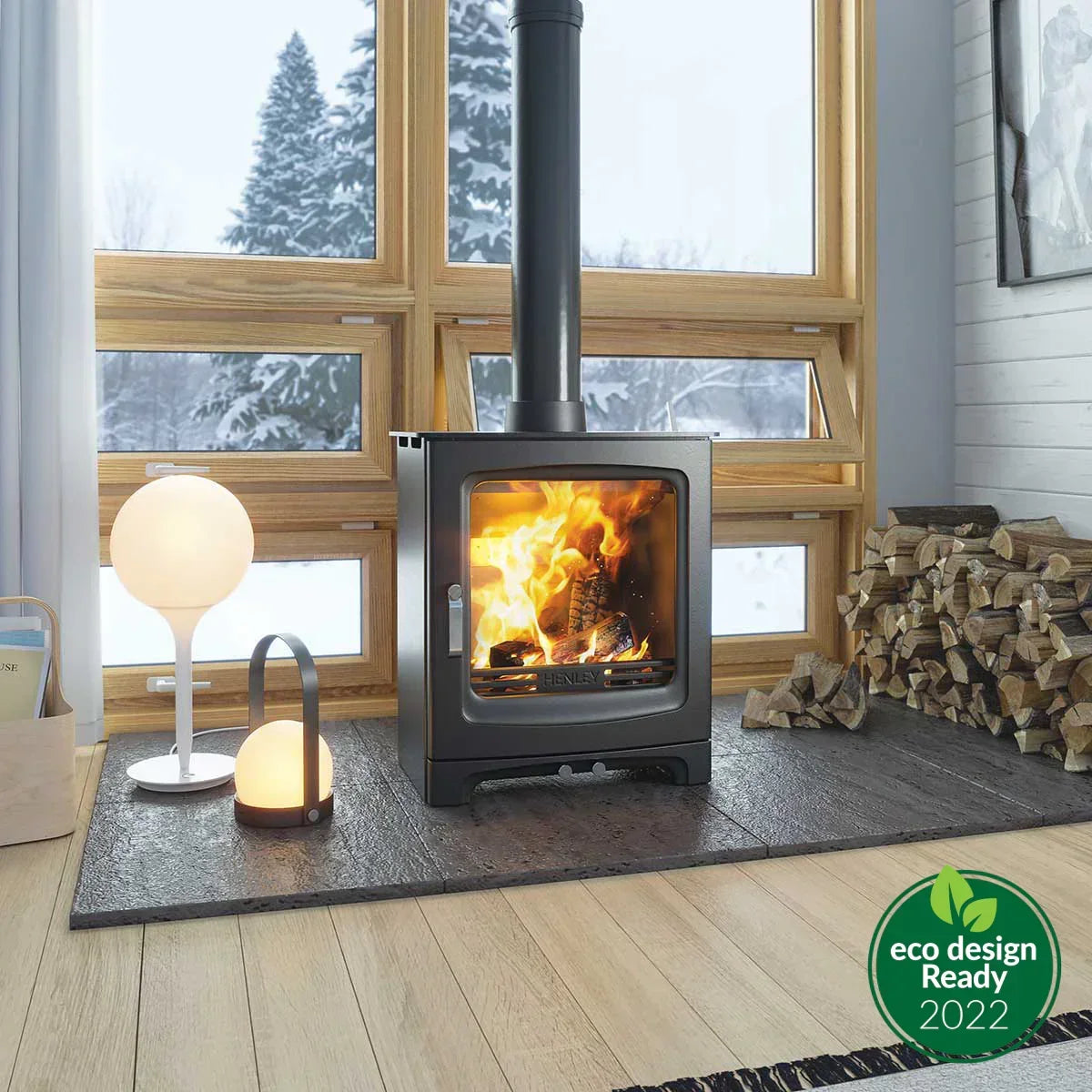 Black wood burning stove with glass door, stacked firewood, and winter scene through window