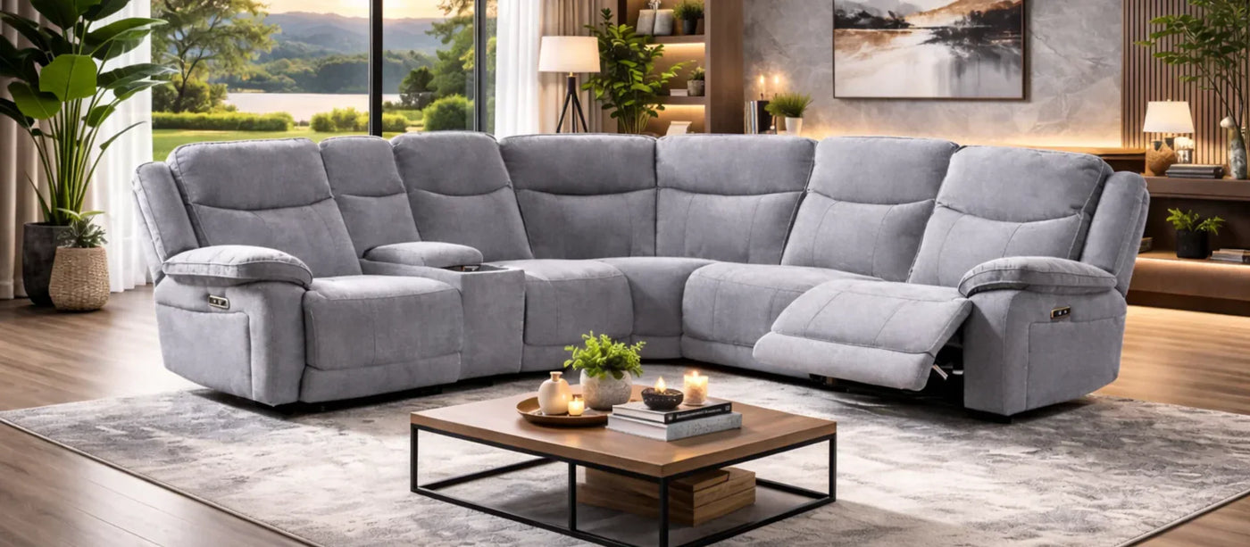A large light gray upholstered sectional sofa with reclining seats, shown in a modern living room setting with a coffee table, rug, plants, and large windows overlooking water.