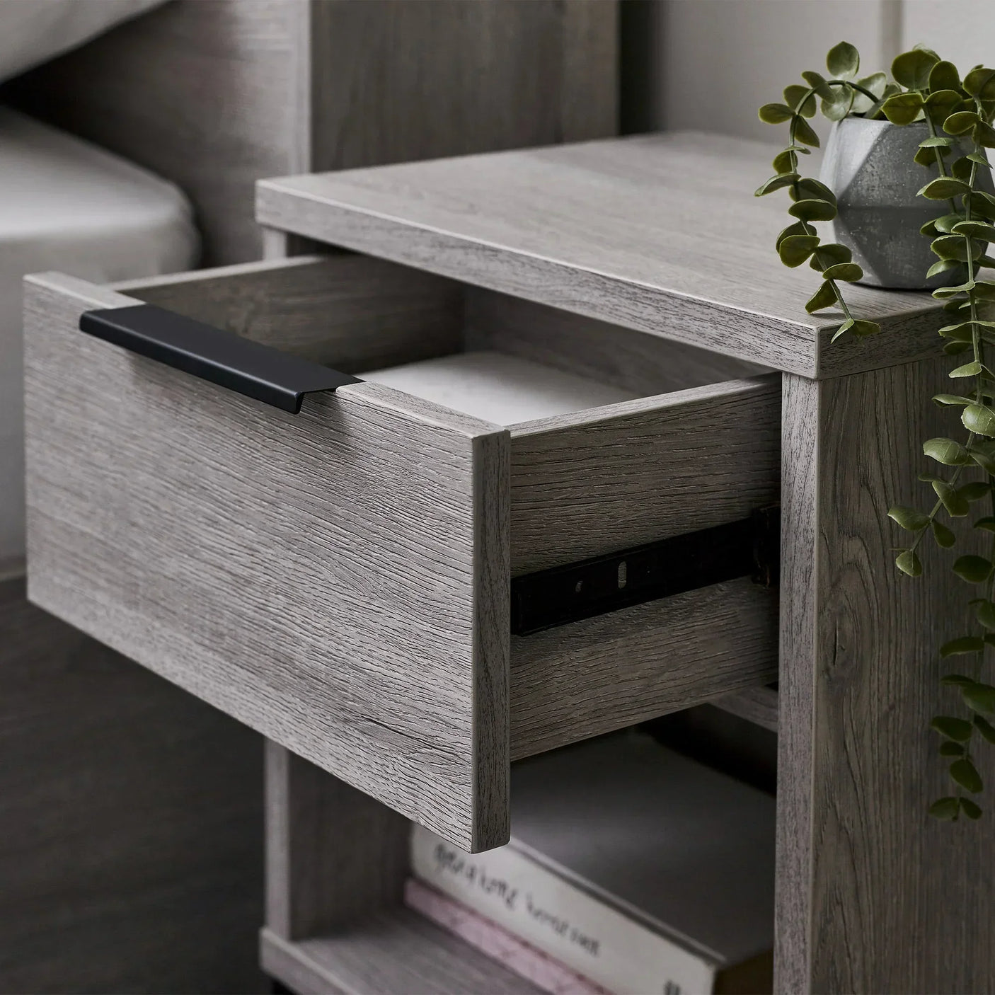 Grey oak bedside table with open drawer, black handle and potted plant on top