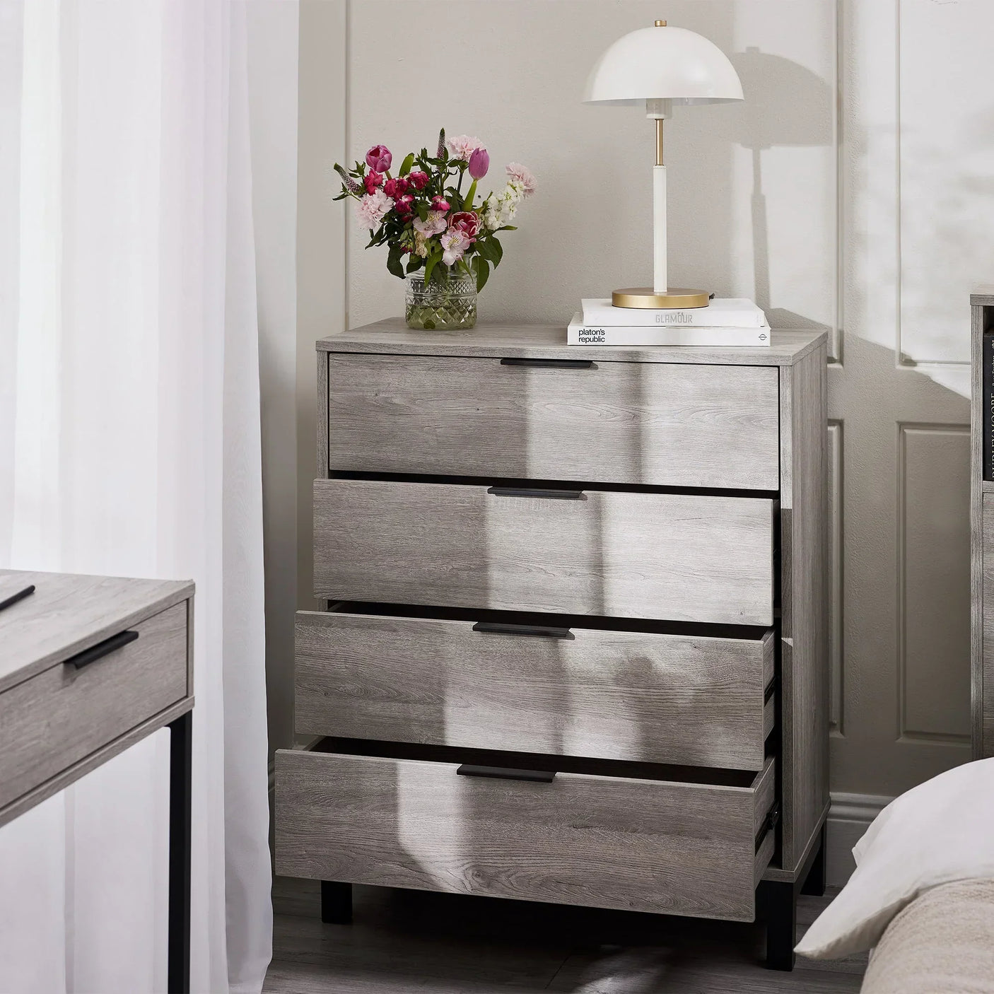 Grey oak 4 drawer chest with black handles, lamp, flowers, and books in modern bedroom