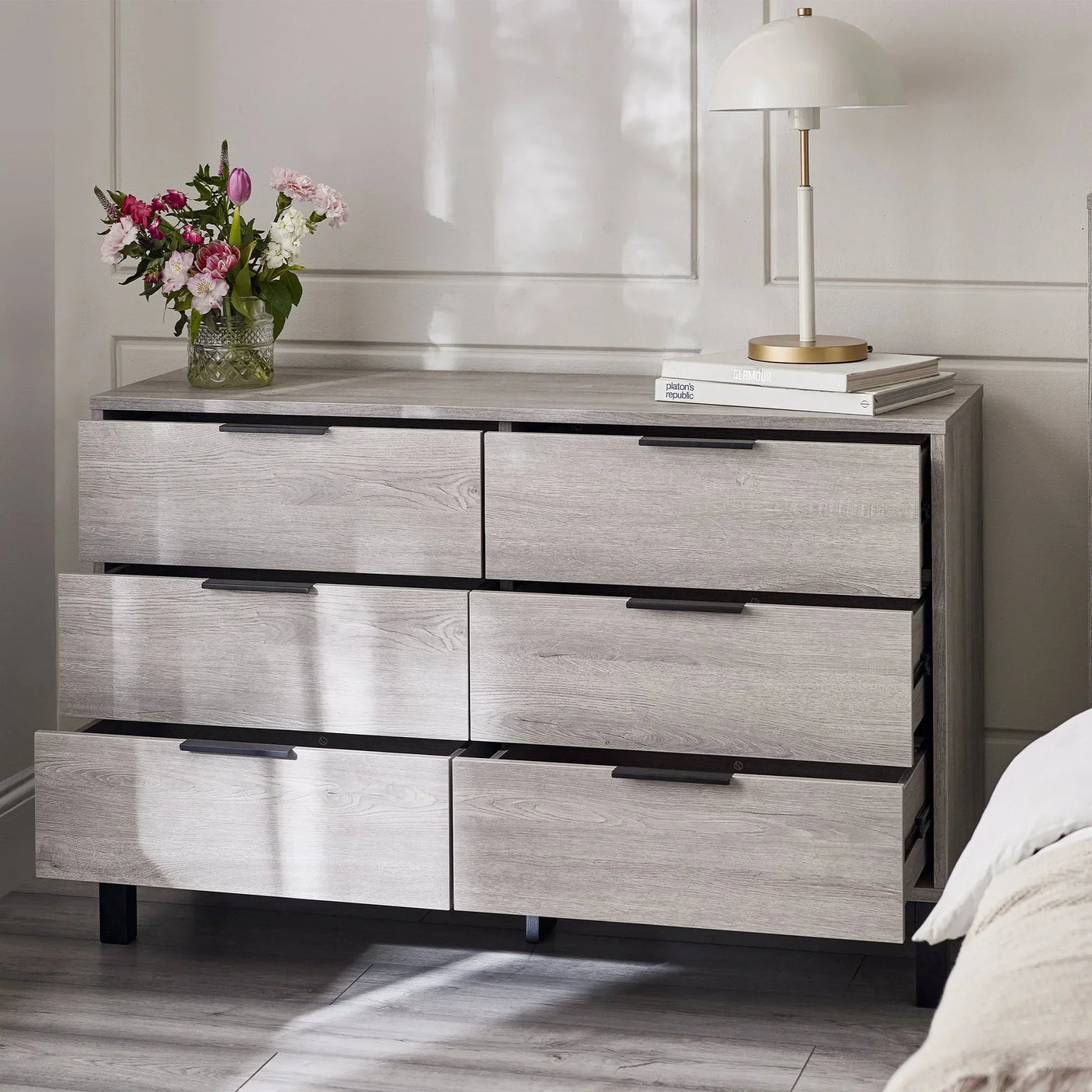 Light grey 6 drawer chest with black handles, styled with lamp and flowers in bedroom setting