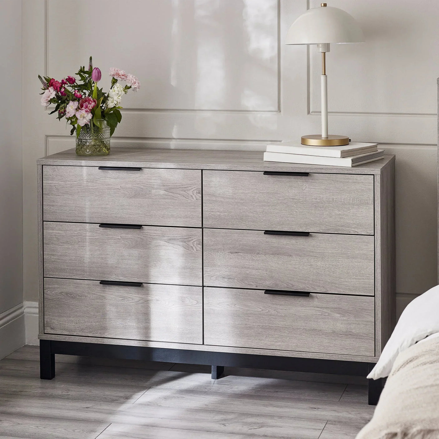 Light wood 6 drawer wide chest with black handles in a bedroom, styled with lamp and flowers