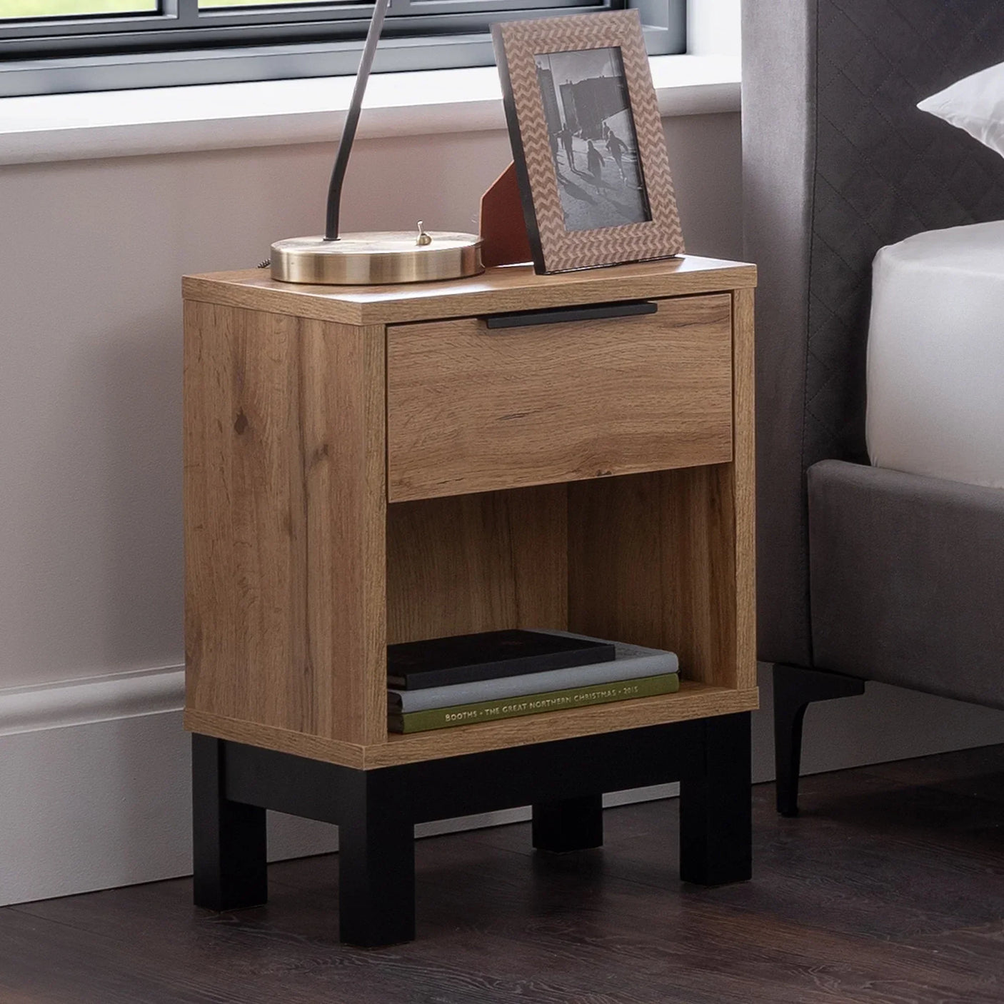 Oak bedside table with one drawer, open shelf, black legs beside bed