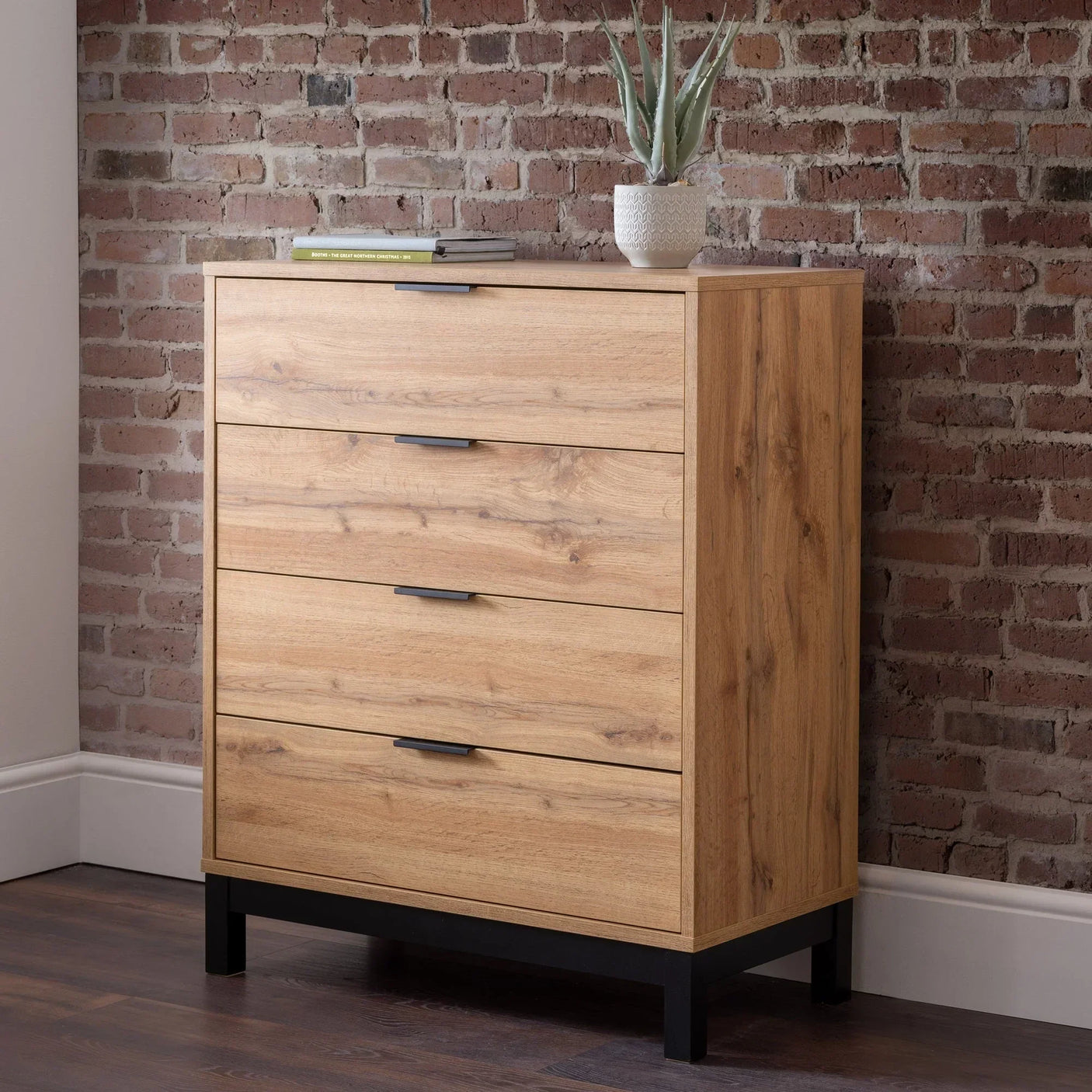 Oak finish 4 drawer chest with black legs against brick wall, plant and books on top