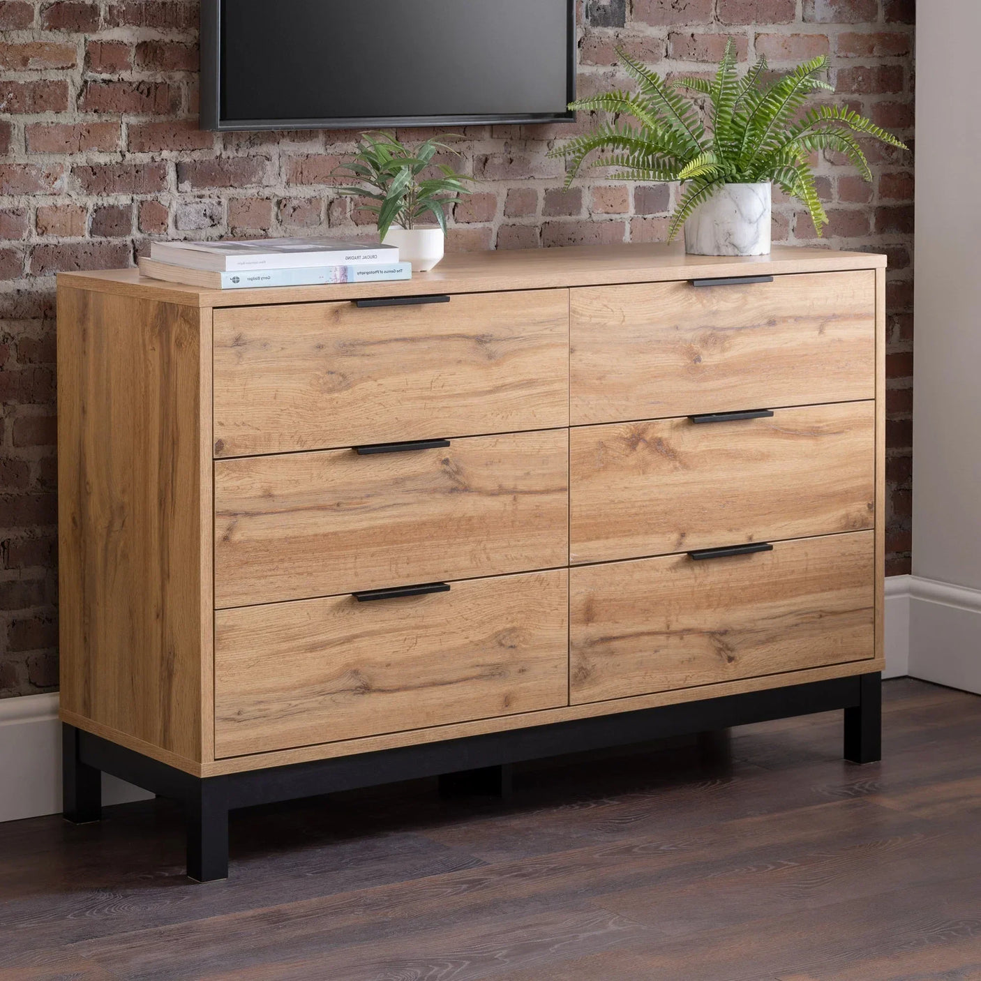 Oak effect 6 drawer chest with black handles, set against brick wall, topped with plants