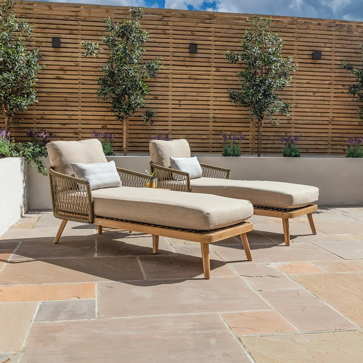 Pair of beige rattan garden loungers with cushions on stone patio with wooden privacy screen