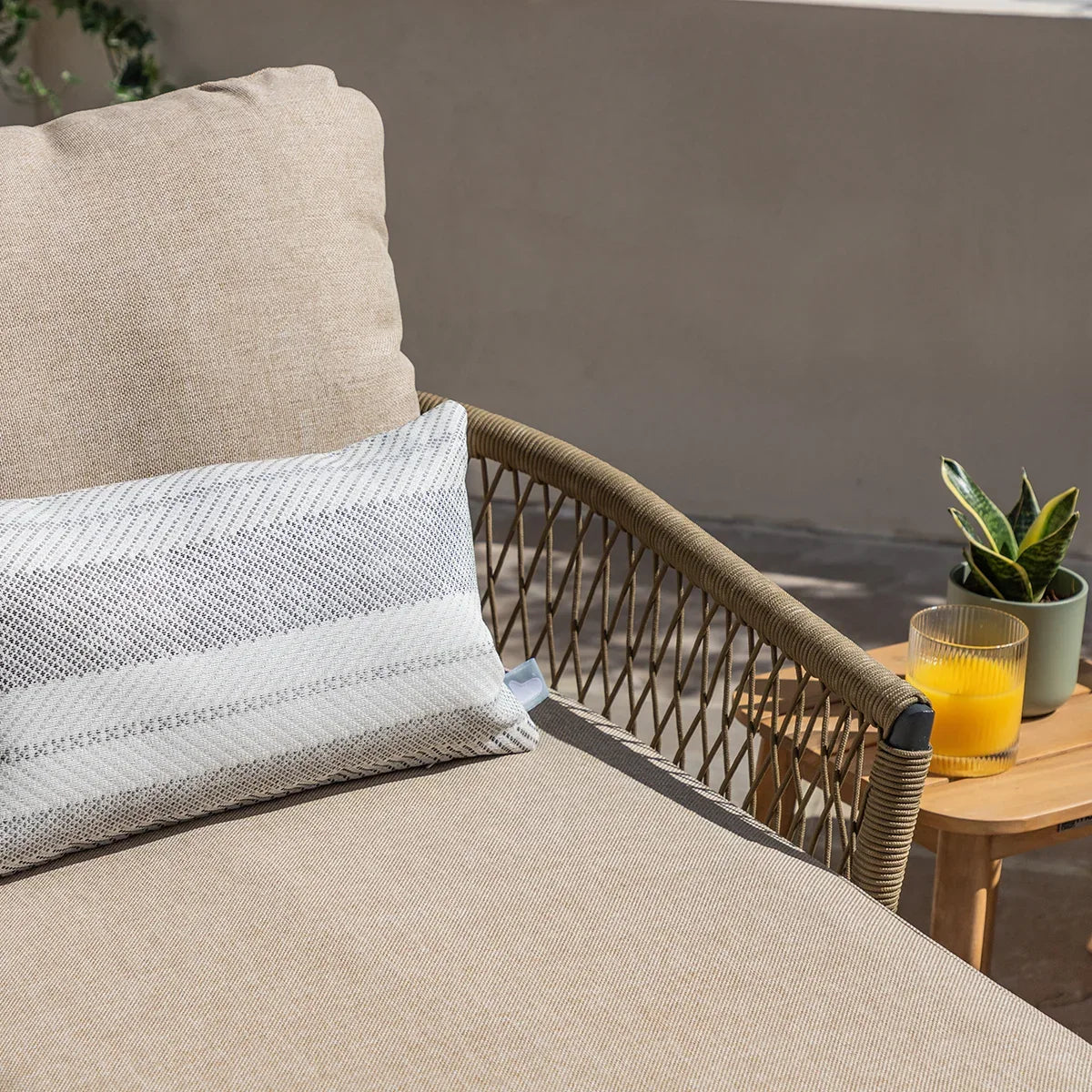 Rattan outdoor sofa with beige cushions and striped pillow beside side table with plant and juice
