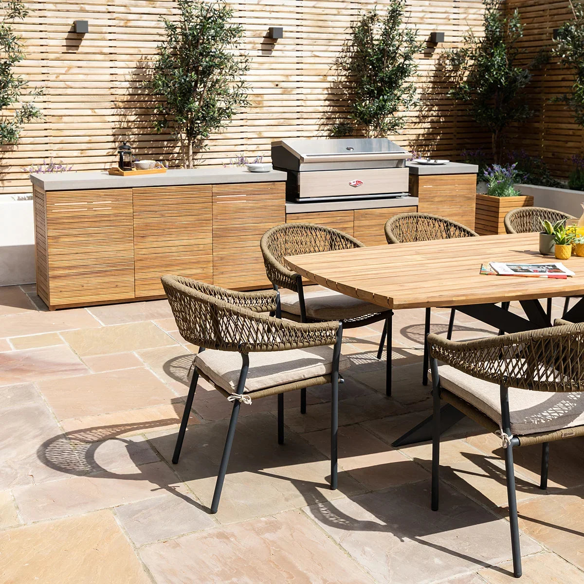 Outdoor dining set with woven chairs and wooden table on stone patio beside outdoor kitchen