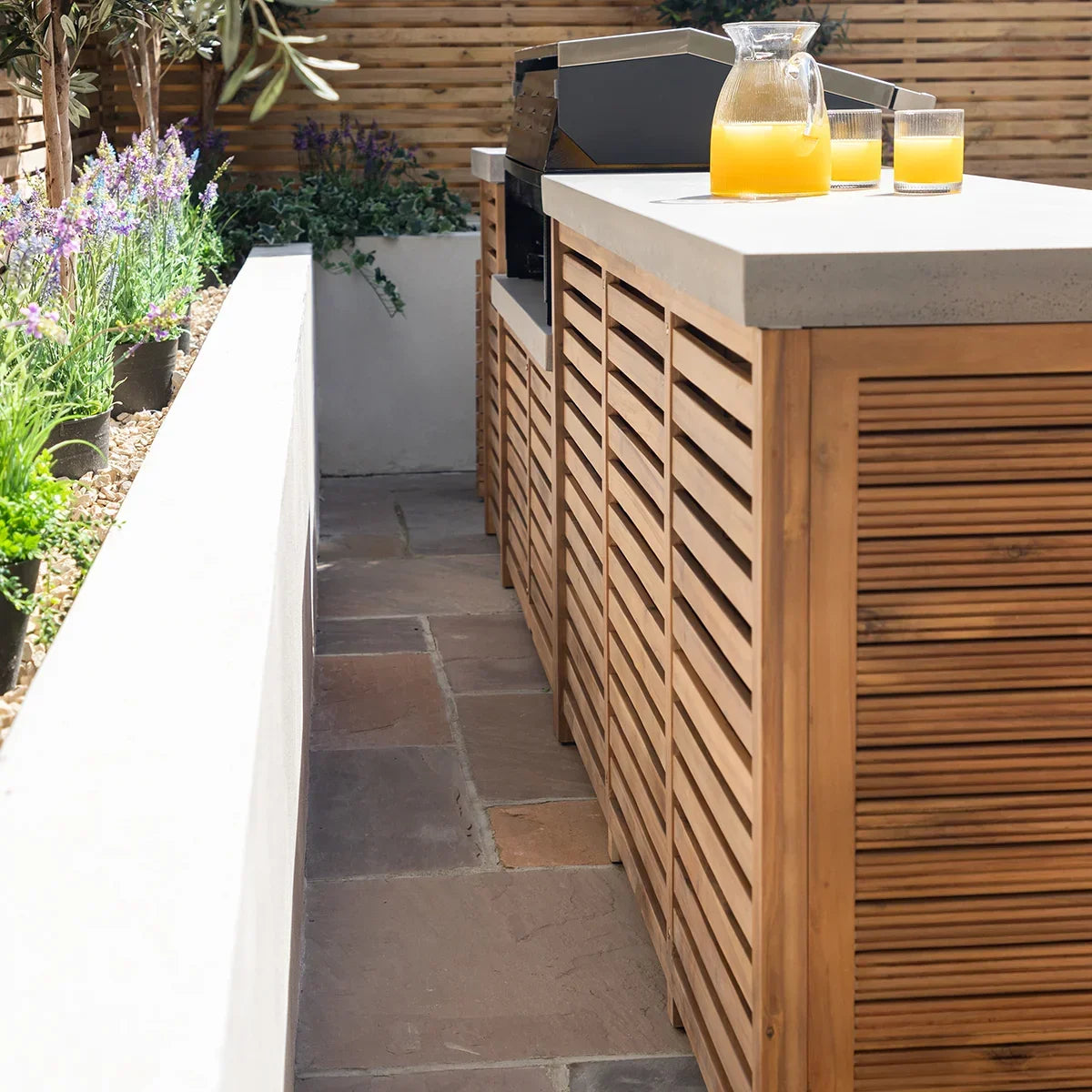 Outdoor wooden kitchen unit with slatted doors and stone top, glasses and jug on surface