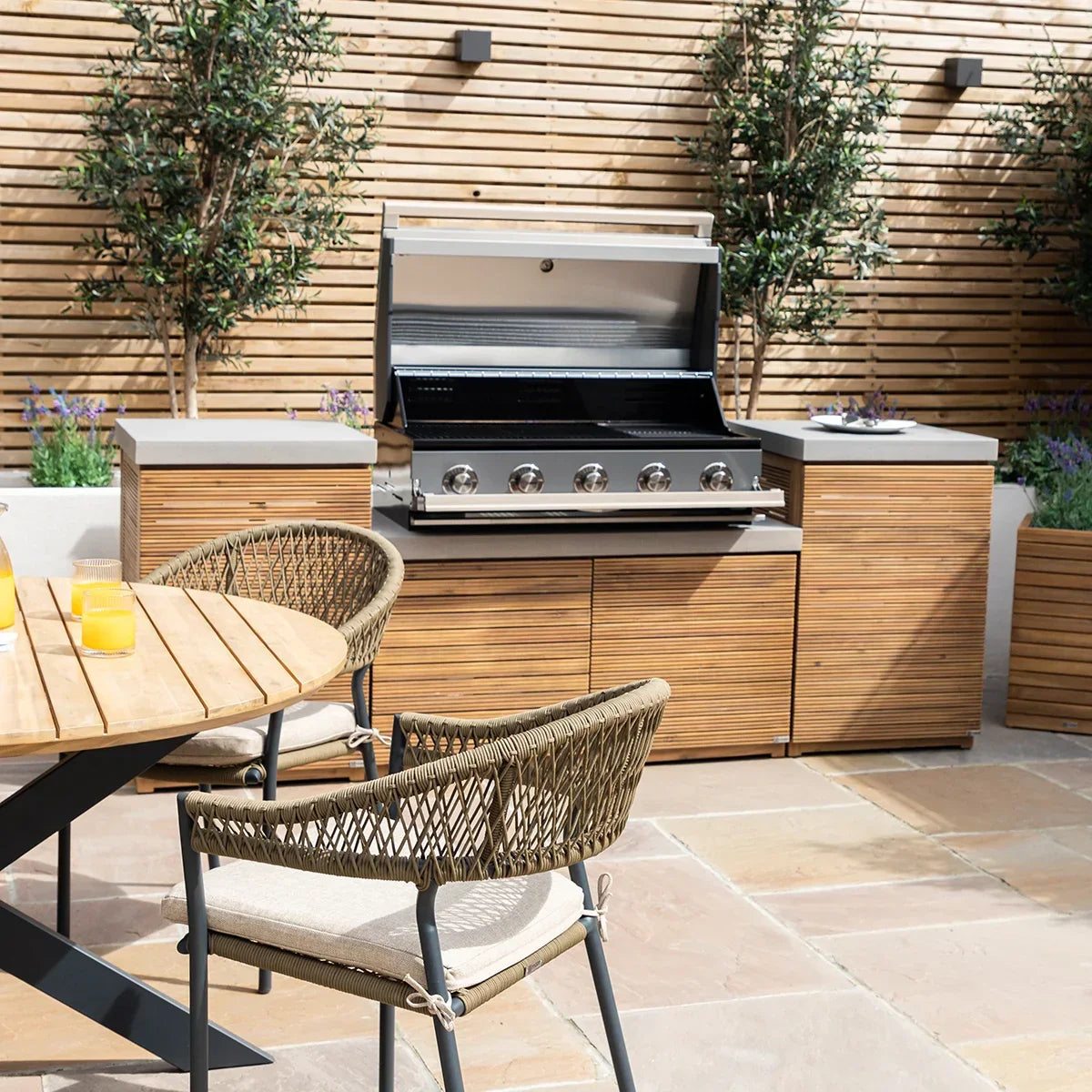 Outdoor wooden dining set with rattan chairs next to built-in gas barbecue and cabinets