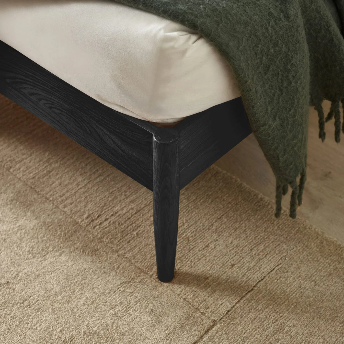Black wood bed frame corner with cream mattress and textured green blanket on beige rug