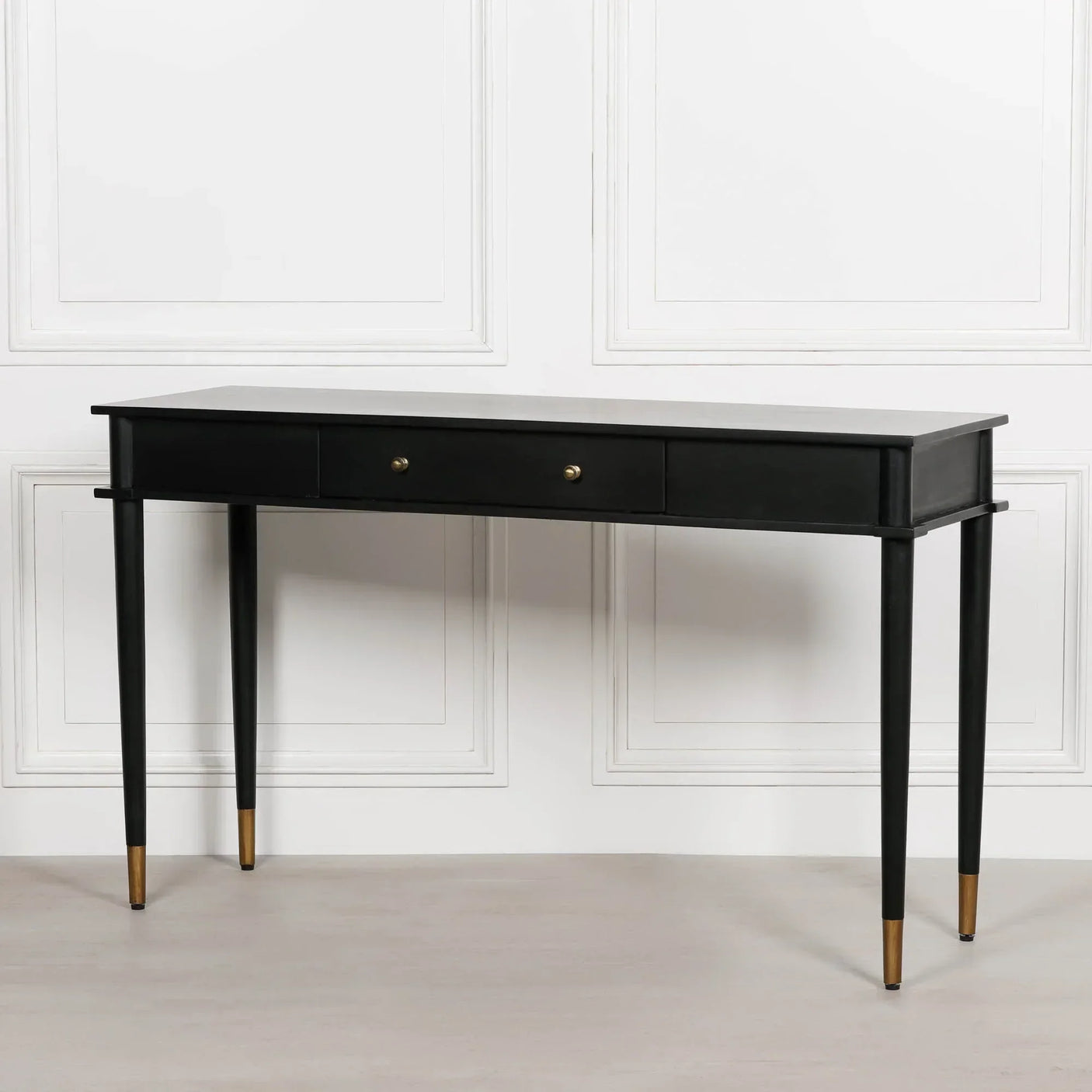 Black console table with two drawers and gold-tipped legs against white panel wall