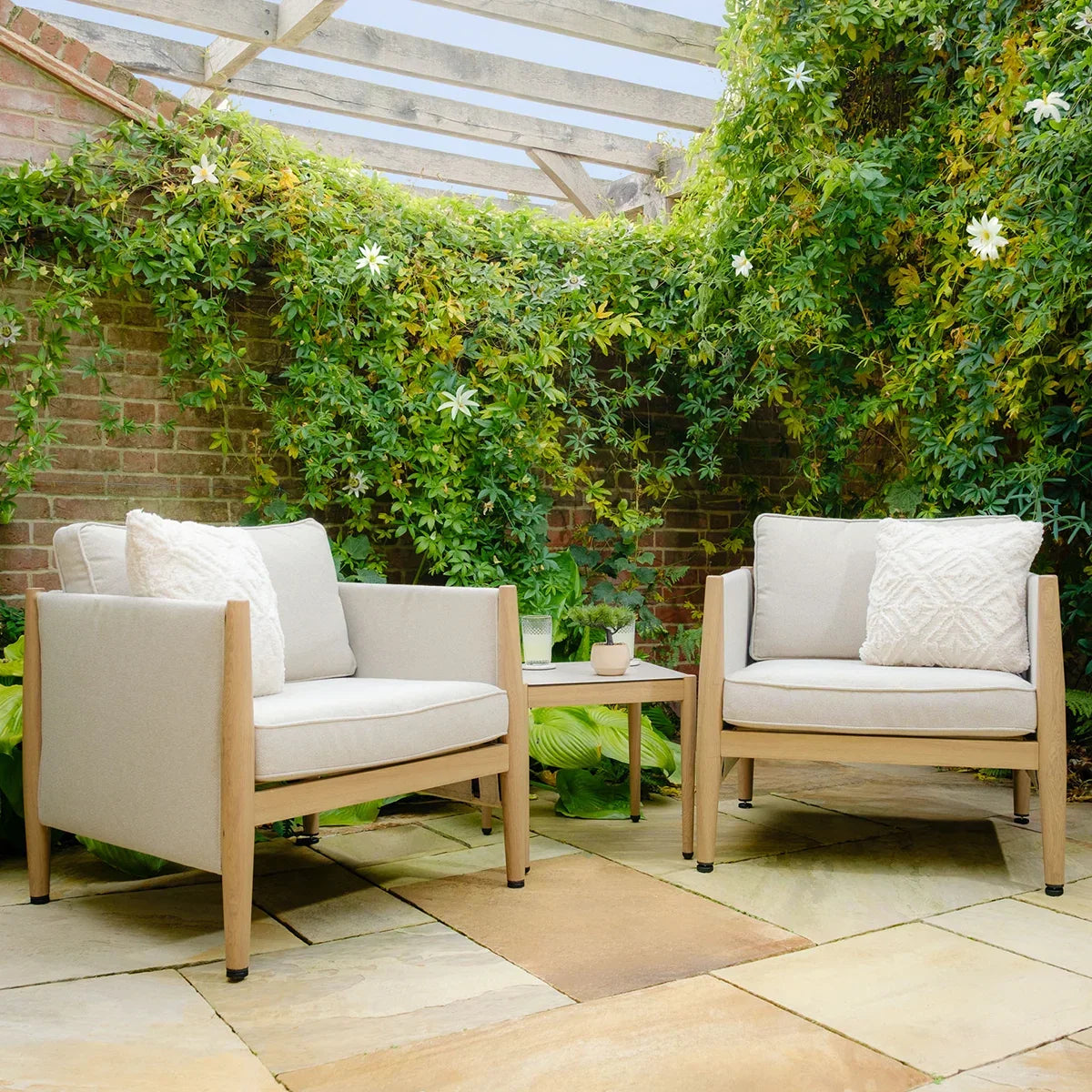 Outdoor lounge chairs with light cushions and side table on stone patio under pergola